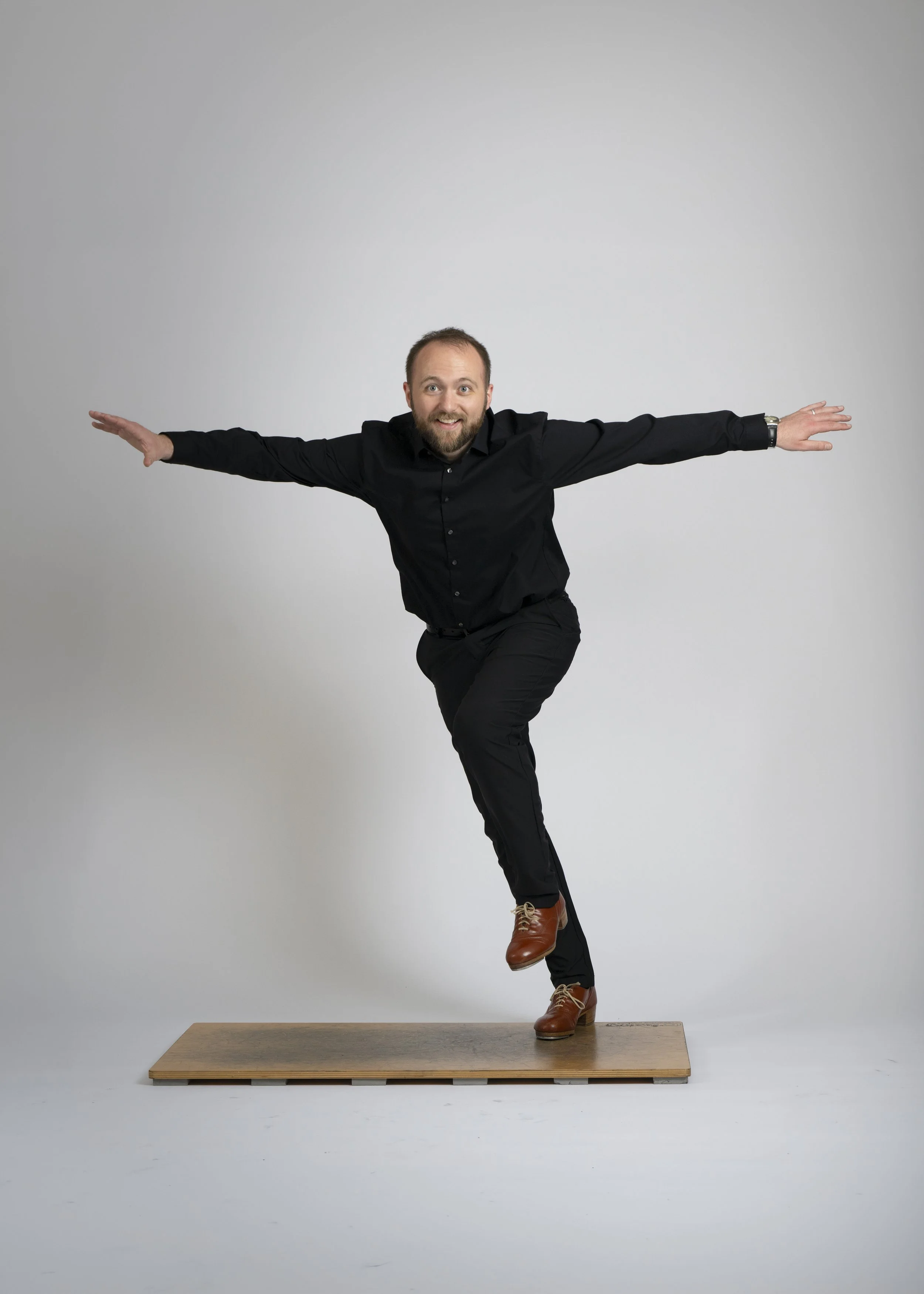 A man with a beard and short hair balancing on one leg on a wooden platform, arms outstretched, wearing a black shirt and brown shoes, against a plain light grey background.