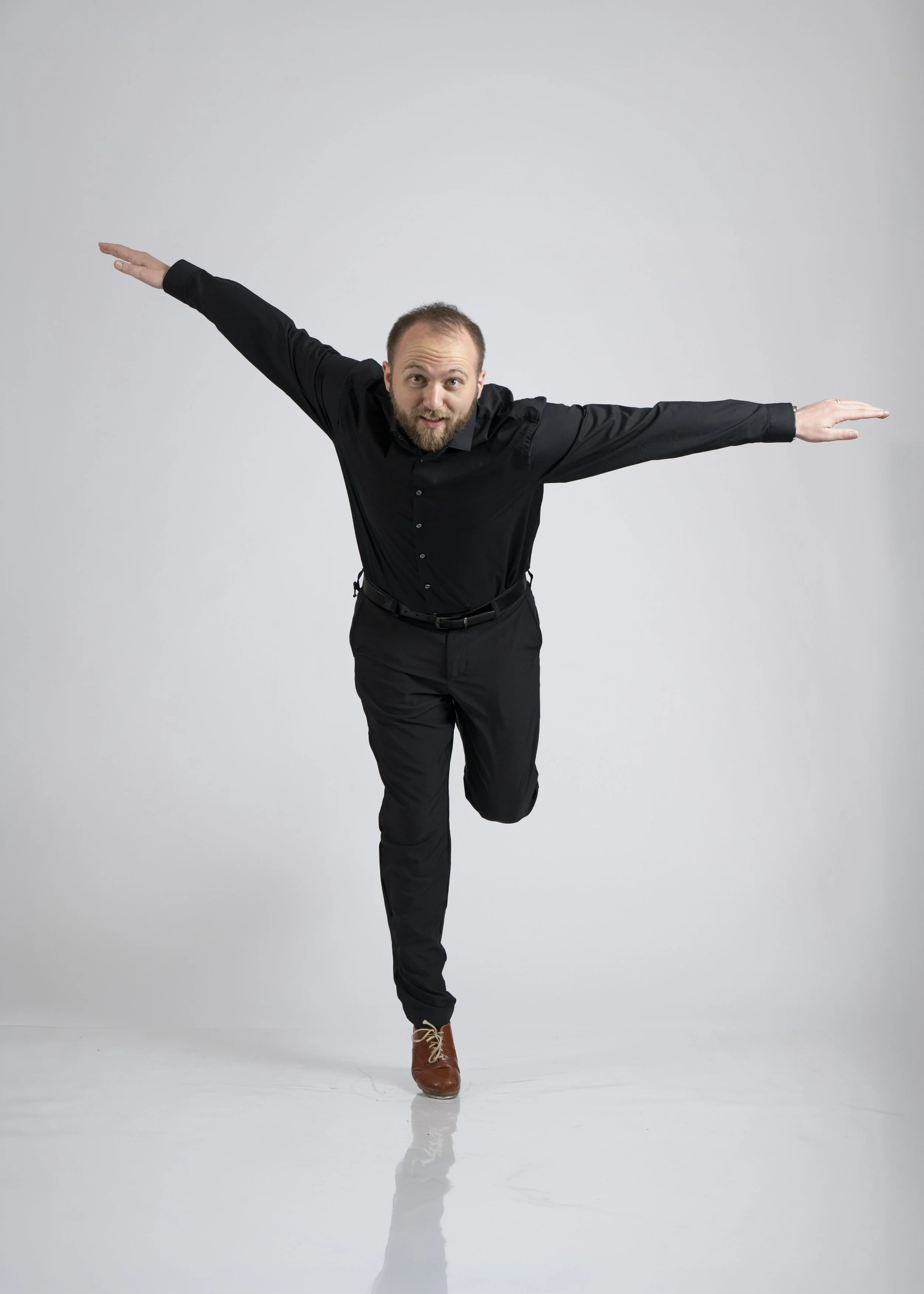 A man with a beard in black clothing, brown boots, balancing on one leg with his arms extended horizontally, against a plain white background.
