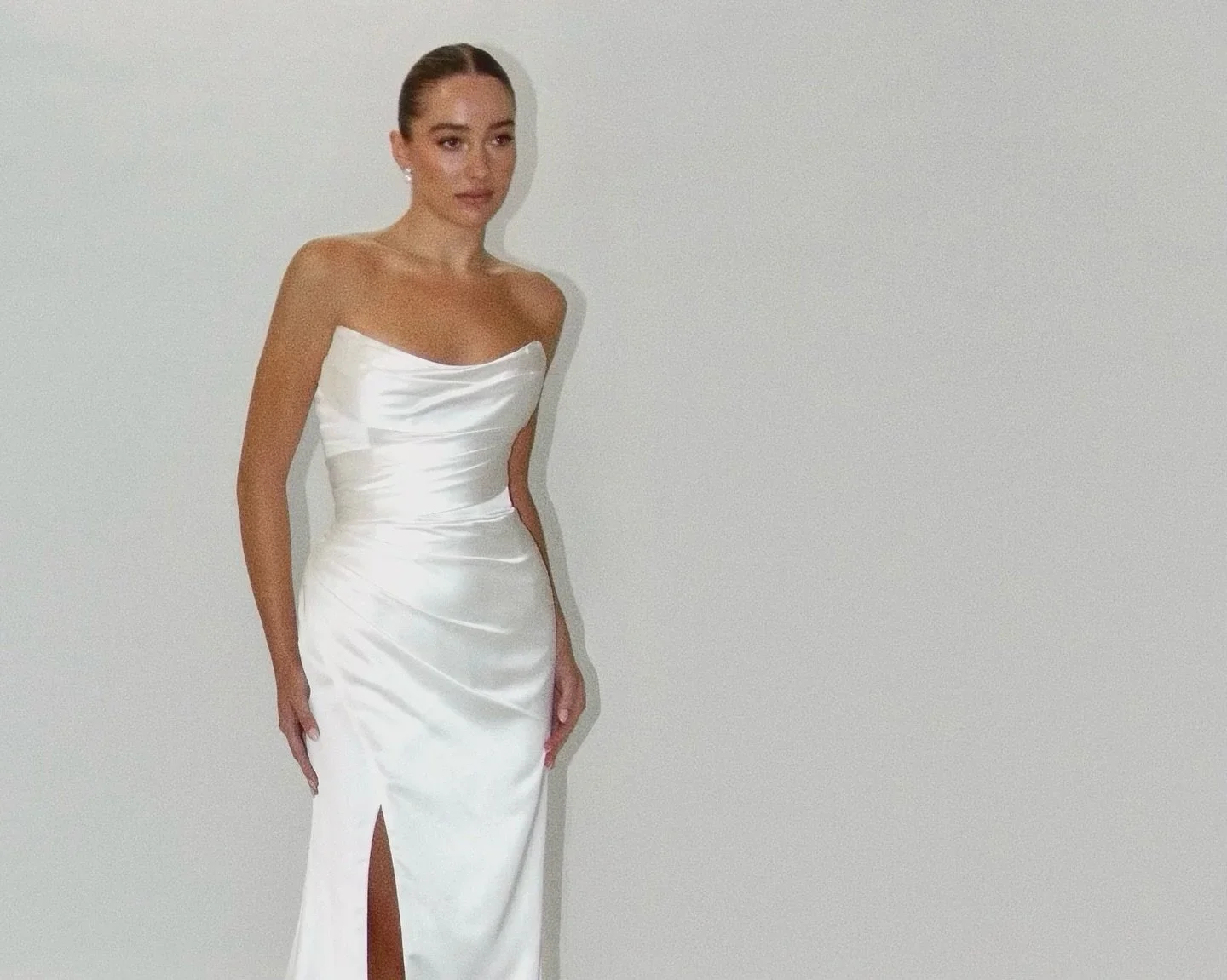 A woman in a strapless white satin gown with a thigh-high slit standing against a plain white wall.