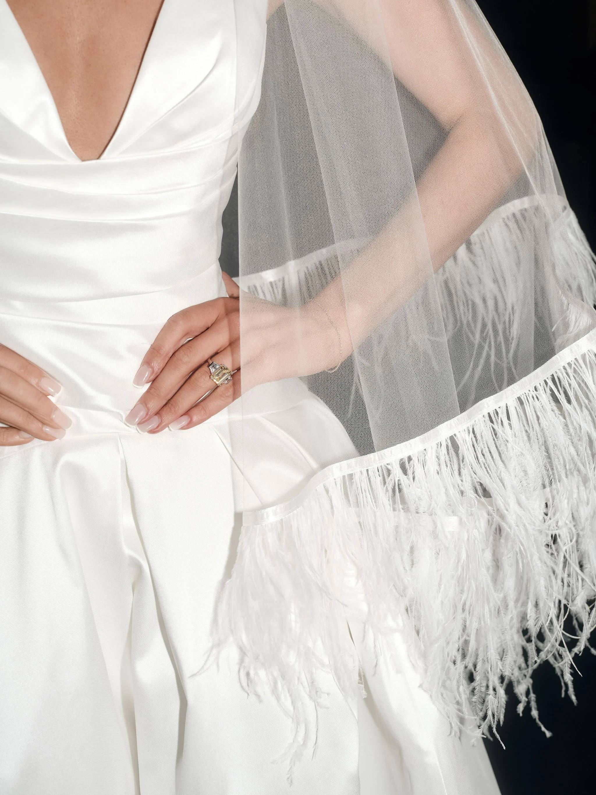 Close-up of a woman dressed in a white satin wedding gown with a deep V neckline, wearing a veil with fringed edges, and a large diamond ring on her left hand.