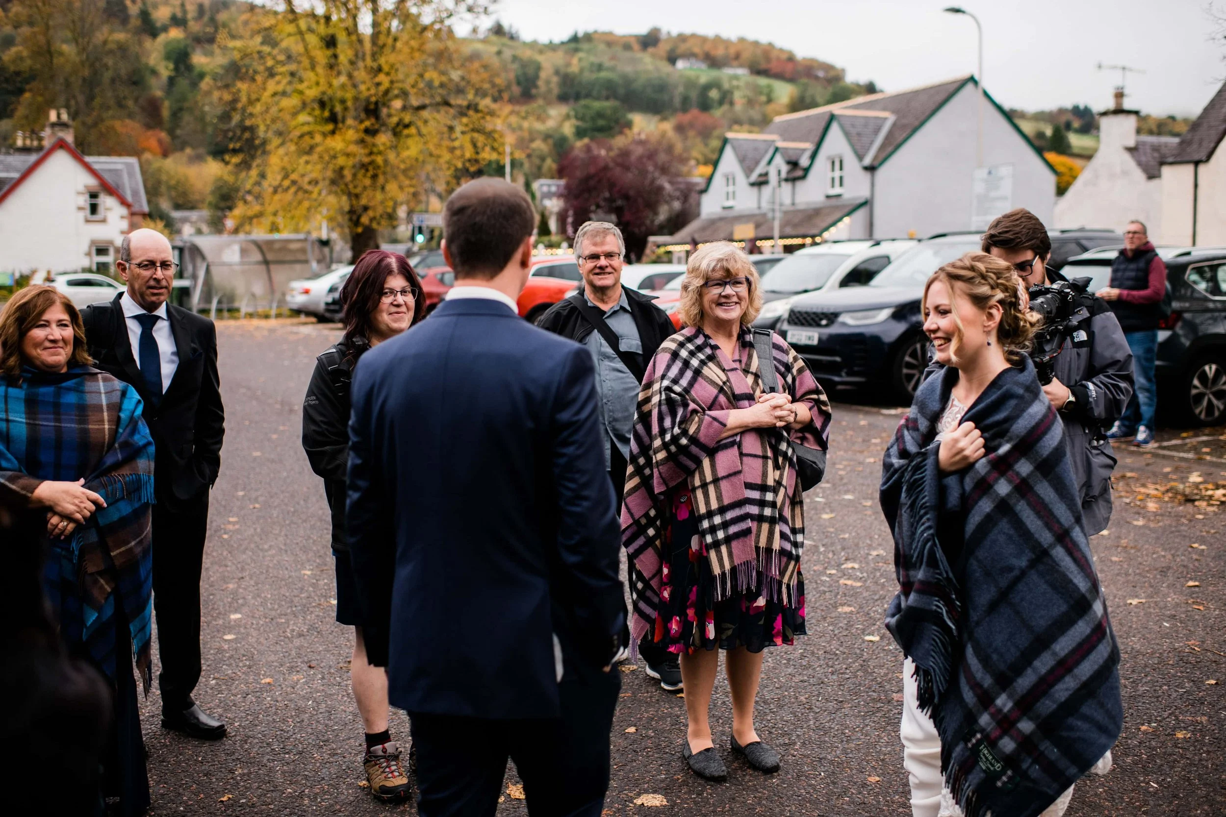 Group of people talking outdoors in fall, some wrapped in plaid blankets, with trees and houses in the background