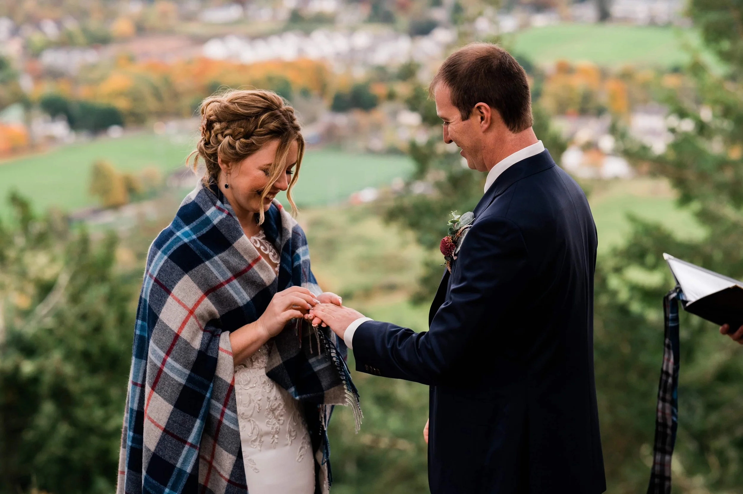 A couple exchanging rings at an outdoor wedding ceremony, with a scenic landscape of green fields and trees in the background, during daytime.