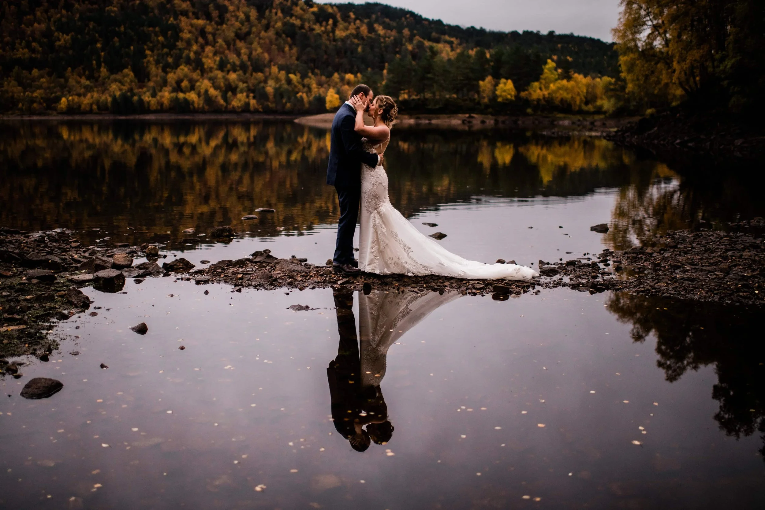 A bride and groom sharing a kiss at the edge of a calm lake during sunset, with autumn trees in the background and their reflection visible in the water.