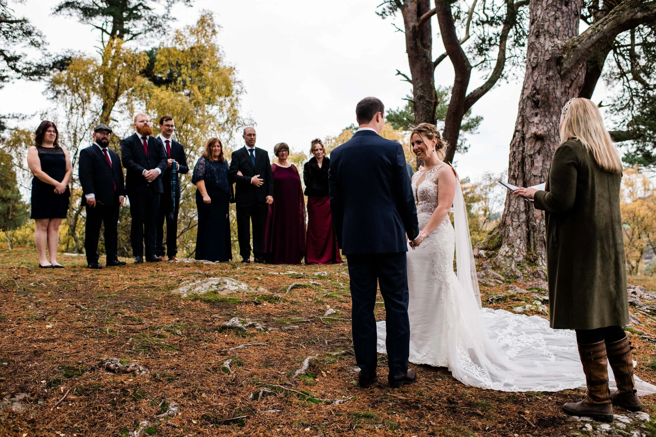 A couple getting married outdoors, holding hands and facing each other, with an officiant reading from a tablet. Guests stand in a line behind them on a forested area with trees and autumn foliage.