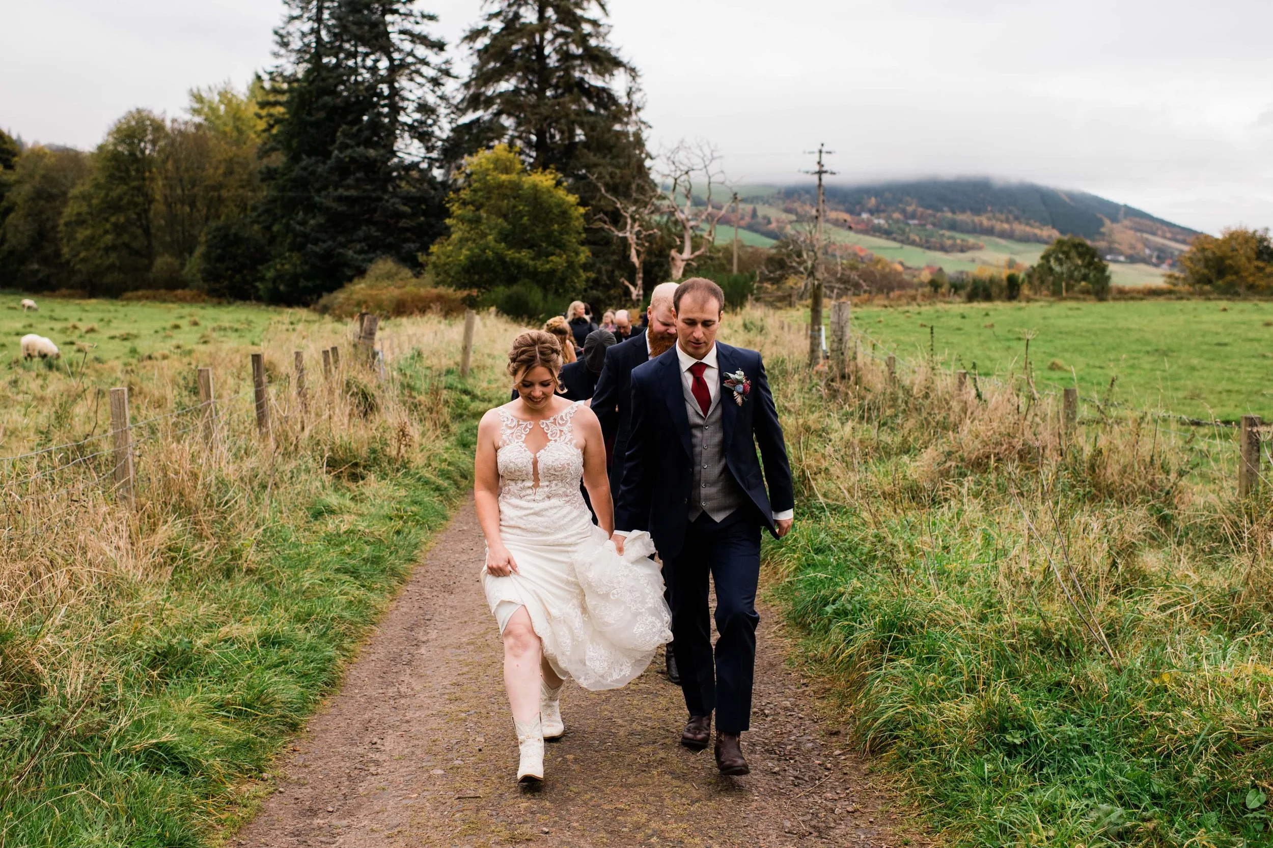 A bride and groom walking along a dirt path in a rural field with friends behind them, overcast sky, trees, and rolling hills in the background.