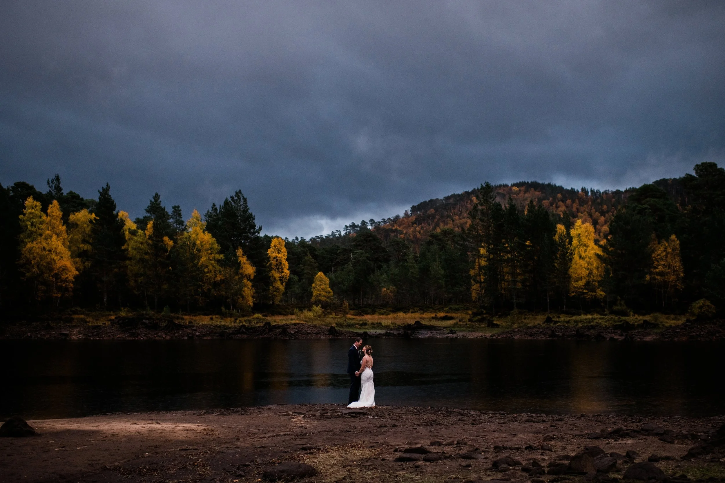A bride and groom holding hands on a beach by a lake at dusk, with trees in autumn colors and dark stormy clouds above.