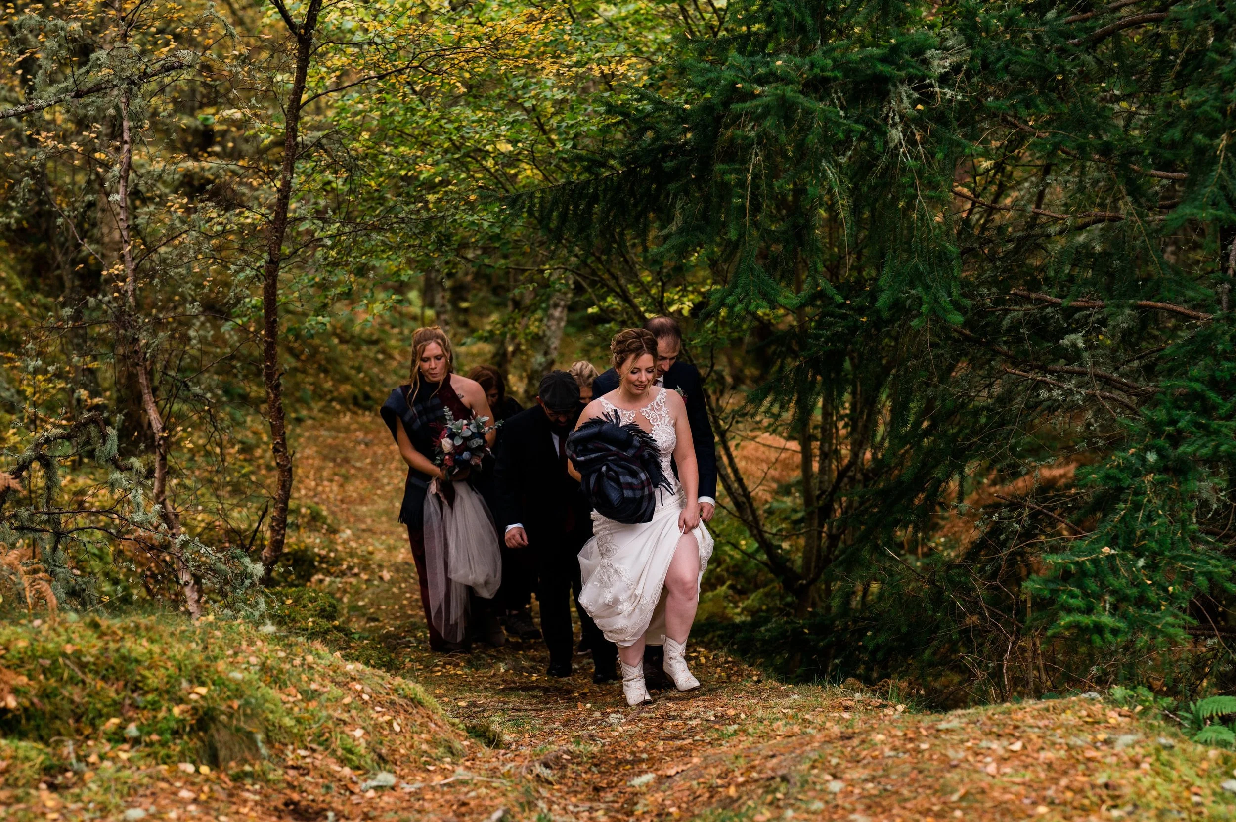 A bride and groom walking through a forested area with wedding guests, some holding bouquets, on a leaf-covered trail.