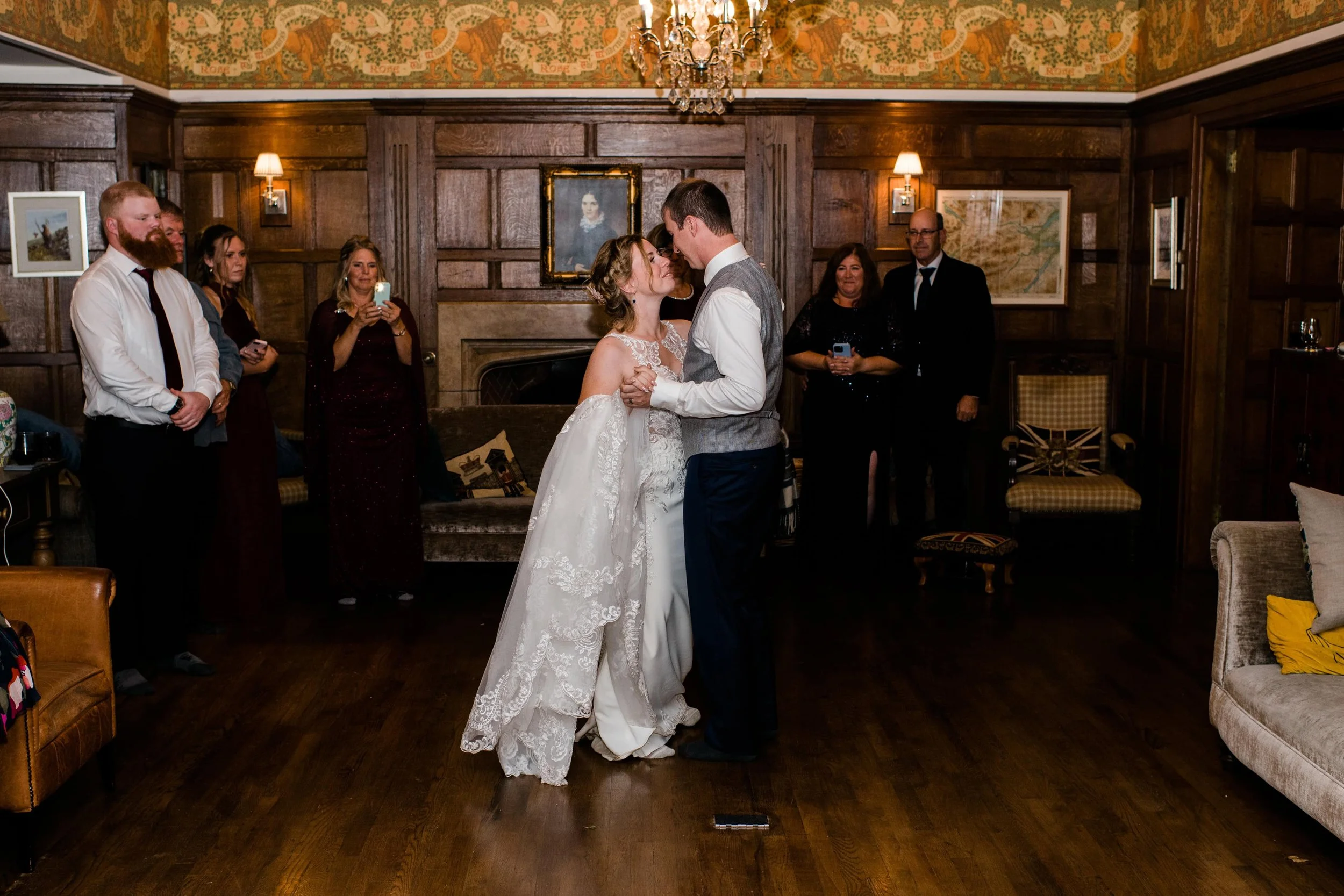 A bride and groom share a dance in a wood-paneled room with family and friends watching and taking photos.
