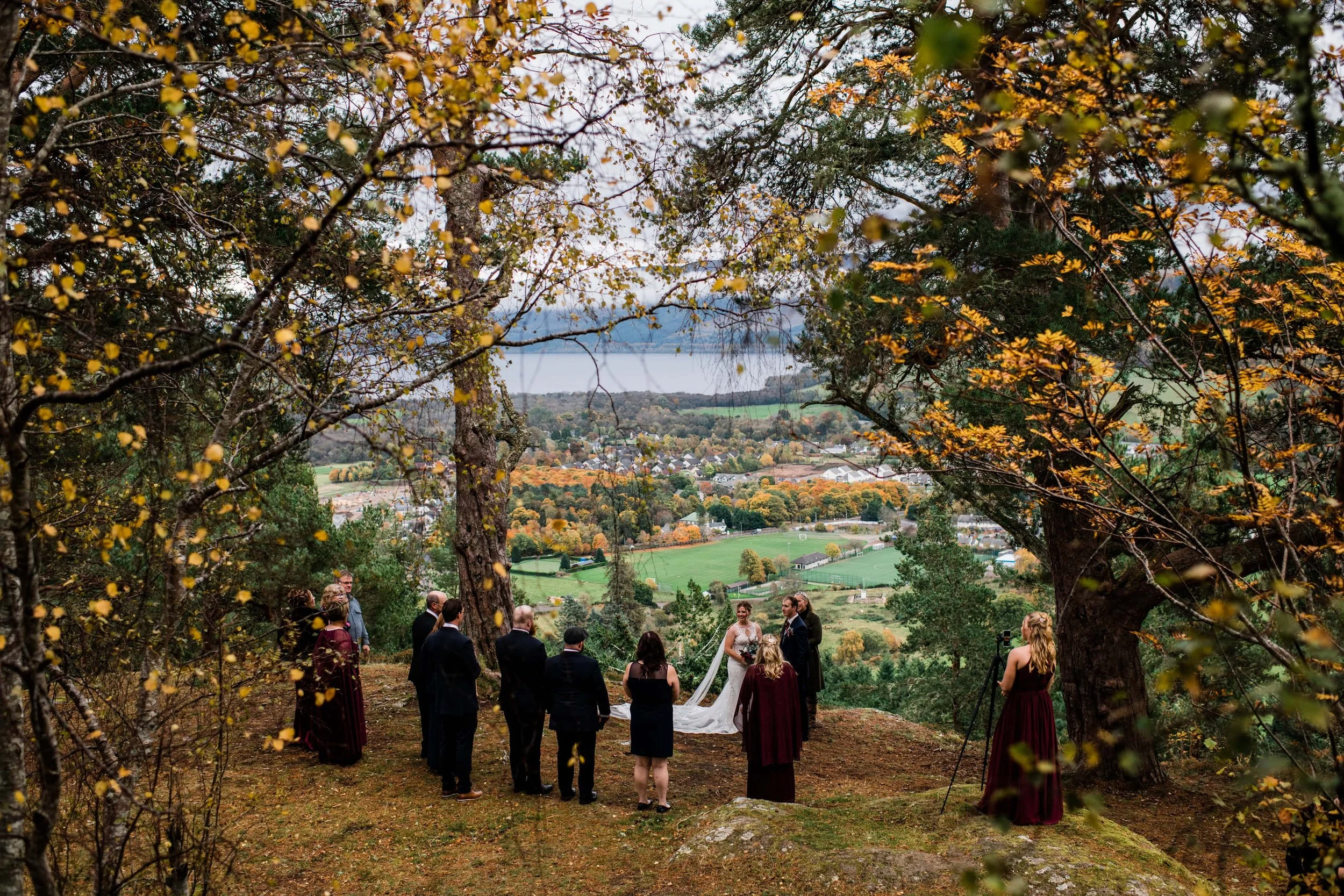 A wedding ceremony taking place outdoors on a hill surrounded by trees with leaves turning yellow and orange. The bride and groom stand in front of their guests with a scenic view of a valley and a lake in the background.