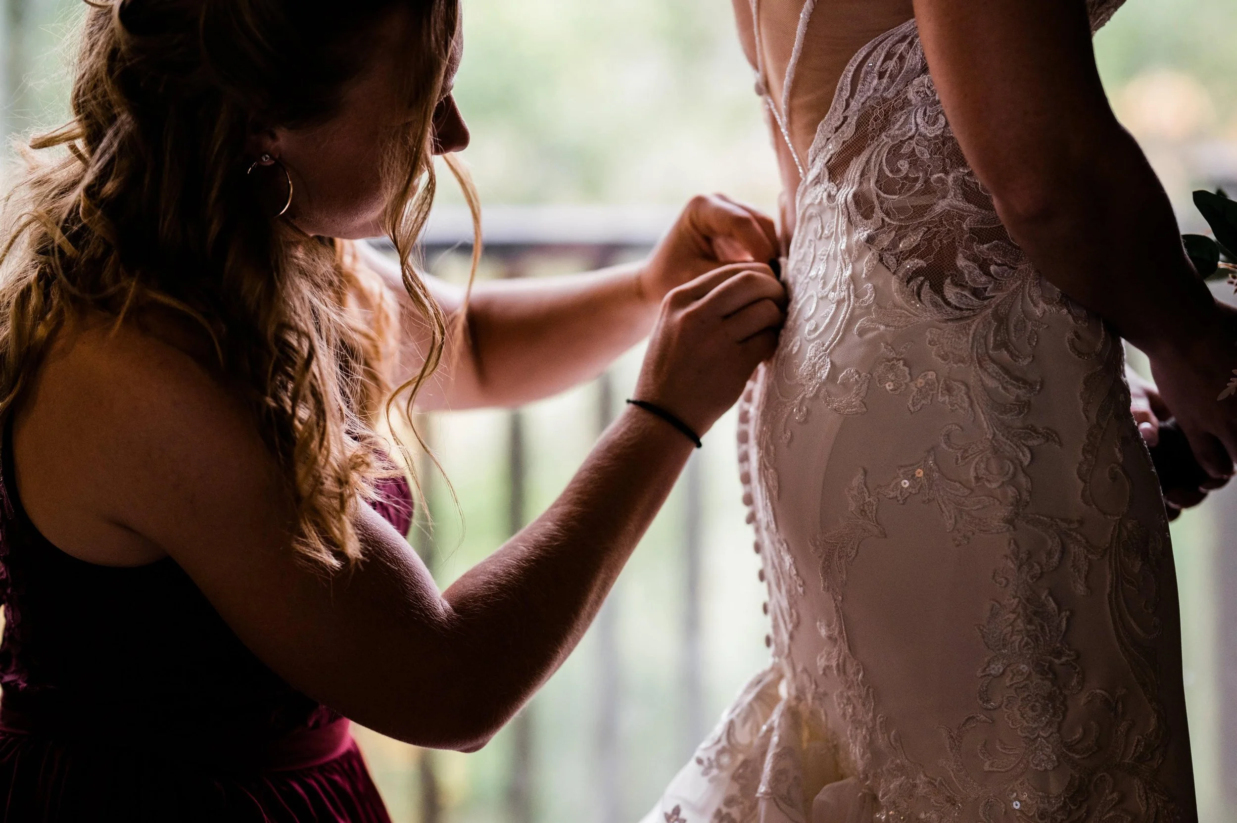 A woman is helping a bride by buttoning her wedding dress, which has intricate lace and embroidery details.