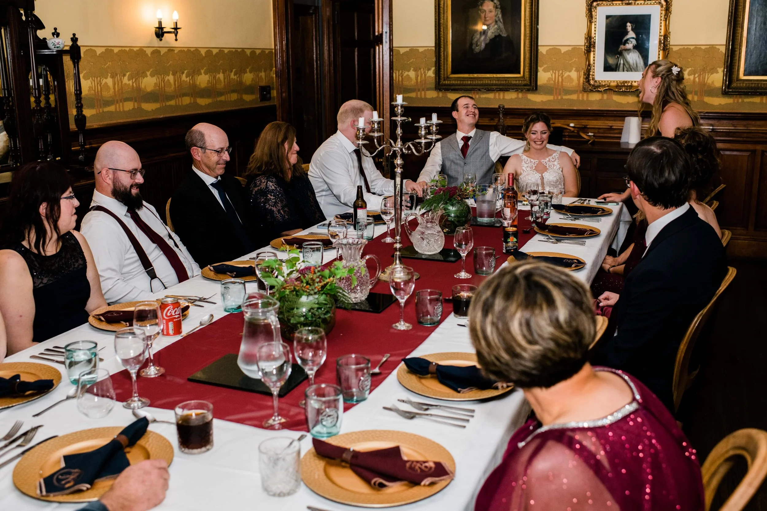 A group of people sitting around a long dinner table, with some of them smiling and engaging with a woman standing at the head of the table, possibly giving a speech or toast. The table is decorated with a red table runner, floral centerpieces, and s