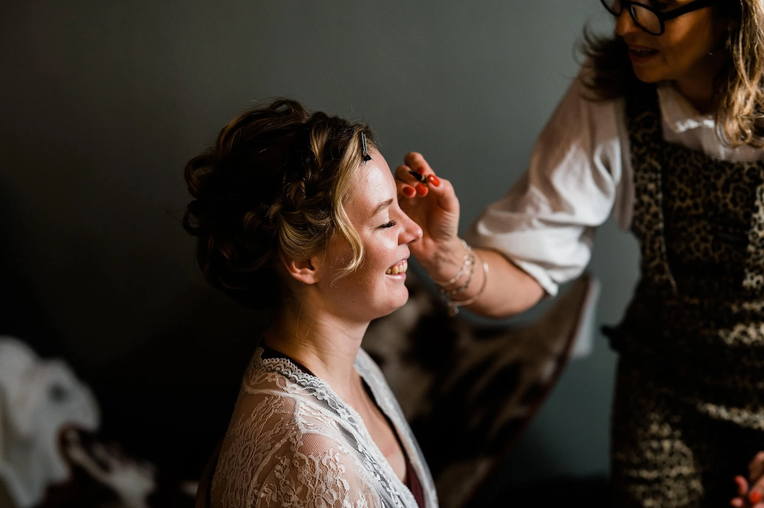 A woman with short curly hair and a lace robe smiling with her eyes closed while another woman, partially visible, applies makeup to her face.