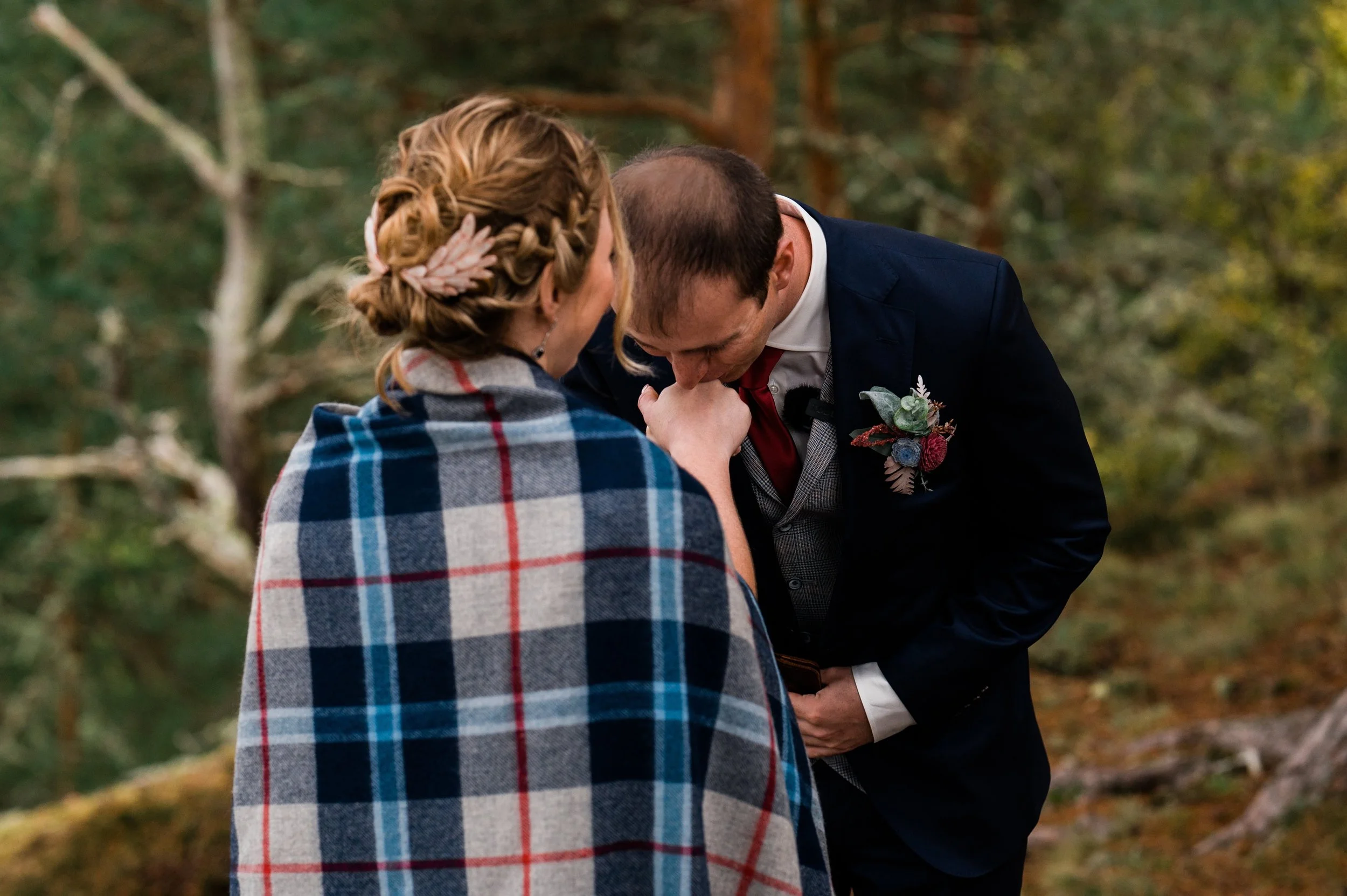 A couple exchanging a kiss outdoors, the man dressed in a dark suit with a boutonniere, and the woman wrapped in a plaid shawl with a leaf hair accessory, surrounded by trees and forest.