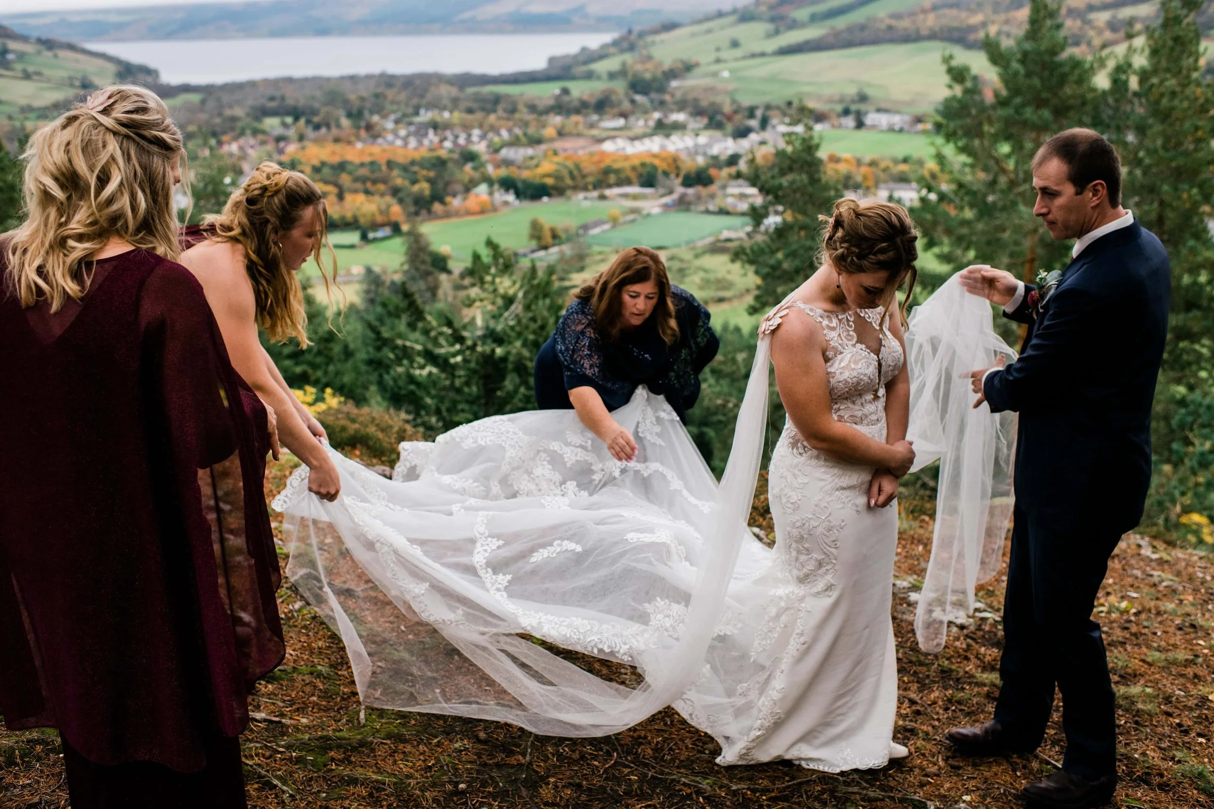 A bride and groom are standing outdoors on a hill with a scenic view of a valley and trees in the background. The bride is wearing a white lace wedding dress, and the groom is in a dark suit. They are surrounded by three women holding the bride's vei
