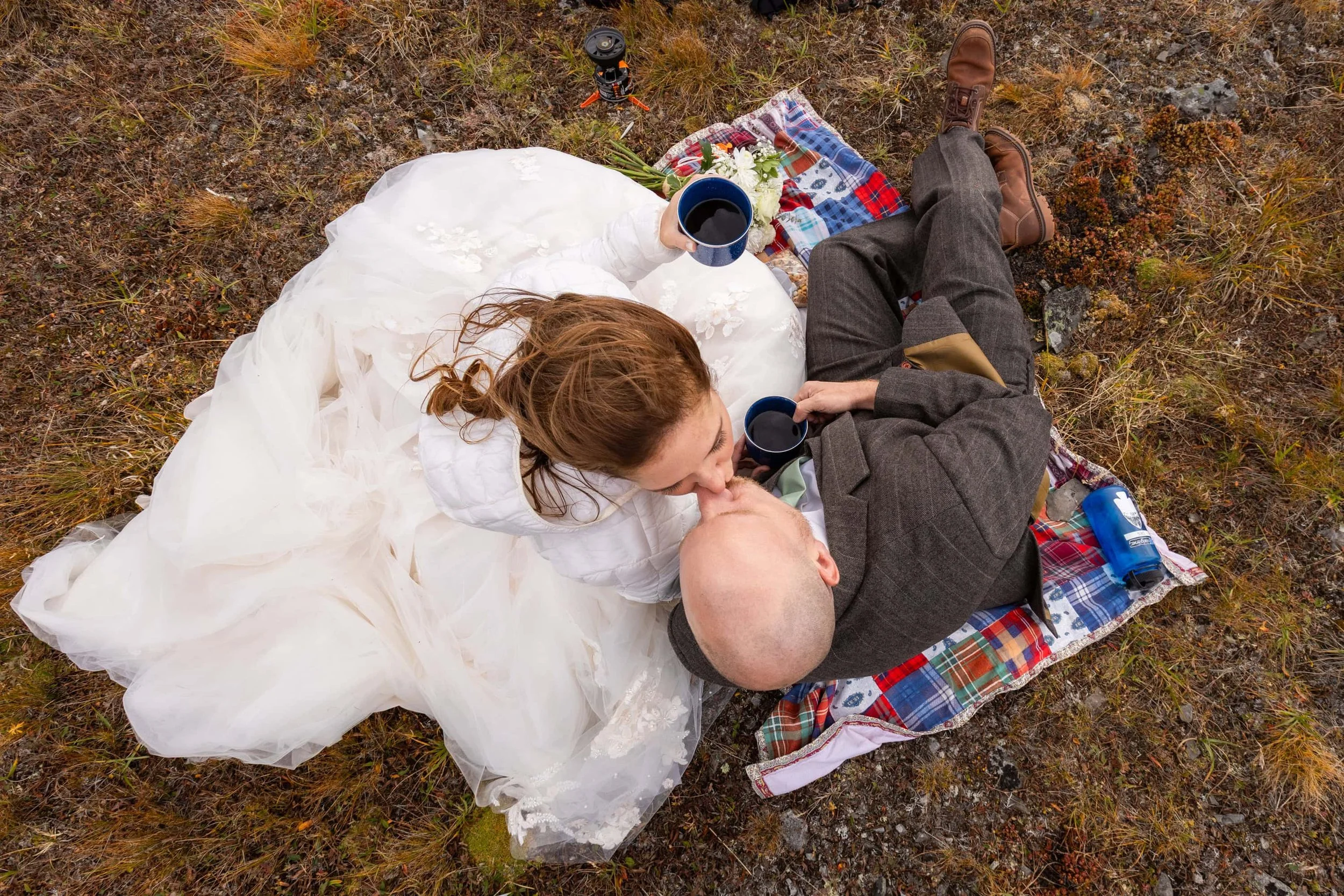 A woman in a white wedding dress kissing a bald man in a dark suit lying on the ground, with two mugs of coffee in their hands. They are on a plaid blanket outdoors in a natural setting with grass and rocks, and there are camping items including a bl