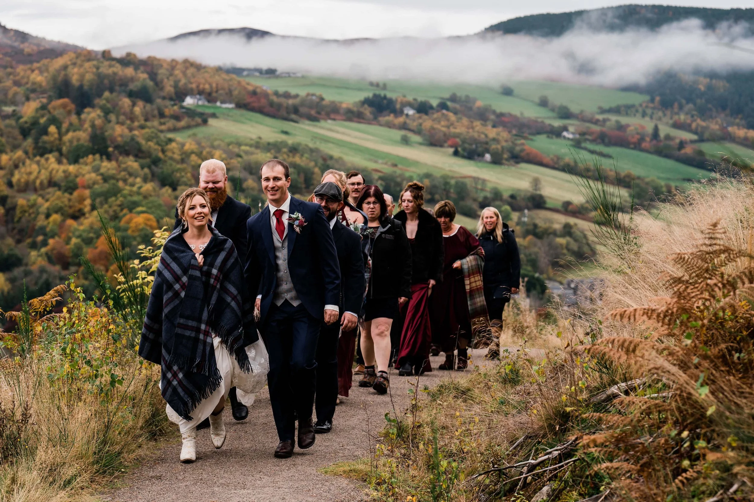 A group of people dressed in formal attire, walking along a scenic hillside trail with autumn foliage and rolling hills in the background.