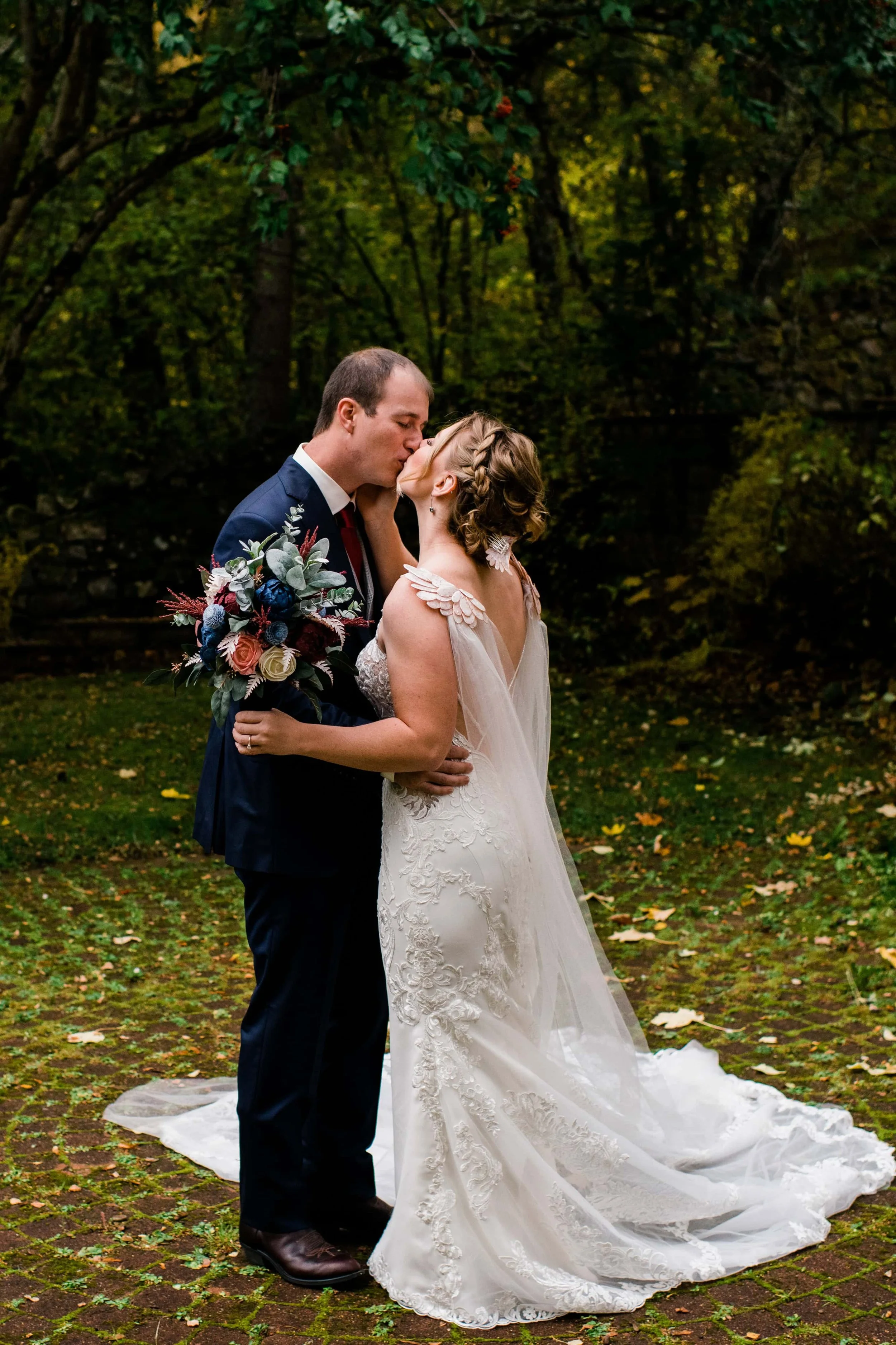 A bride and groom sharing a kiss outdoors on a wooded area, with the bride holding a bouquet of flowers.