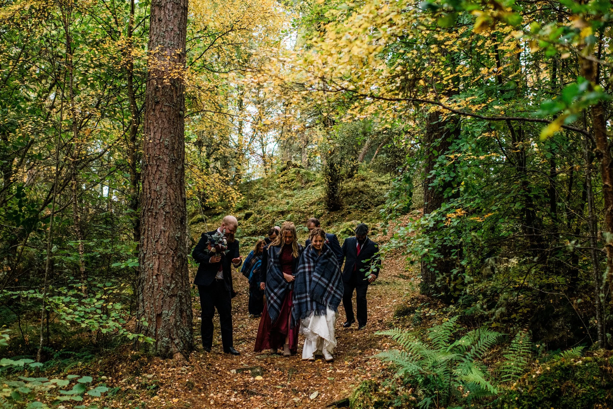 Group of people dressed in formal attire, some wrapped in plaid blankets, walking on a wooded forest trail during autumn.