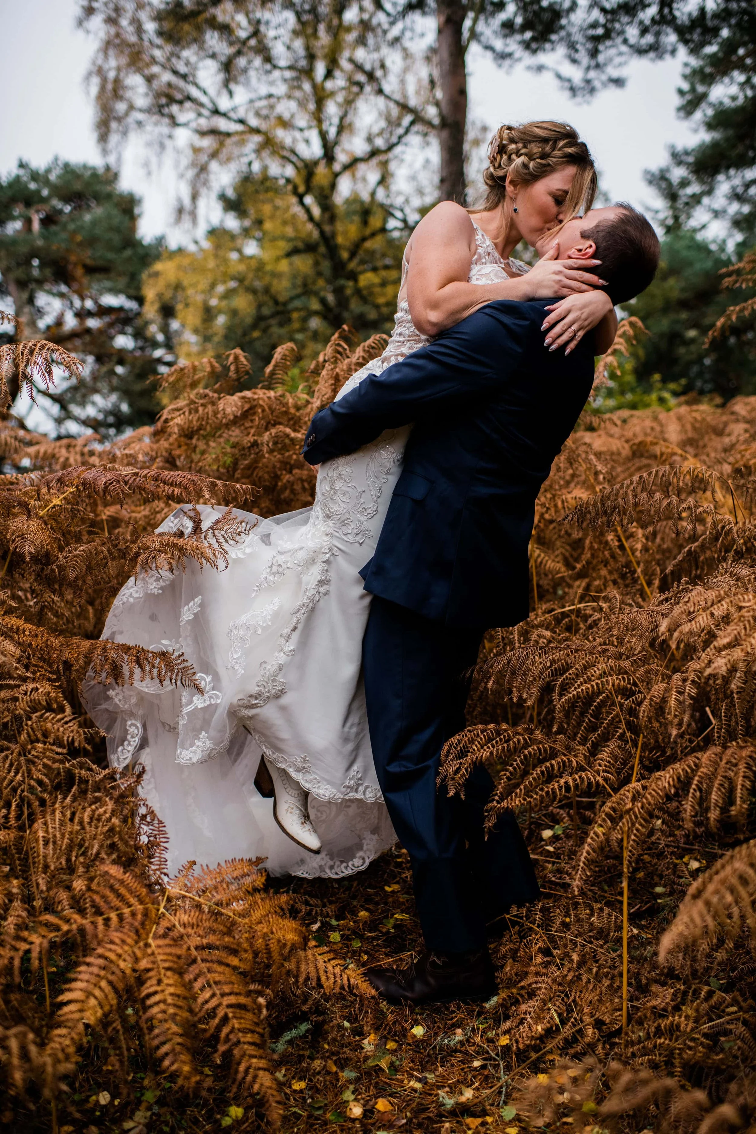 A newlywed couple sharing a kiss in a forested area with autumn foliage. The groom is lifting the bride, who is wearing a white lace wedding dress, while surrounded by brown ferns.