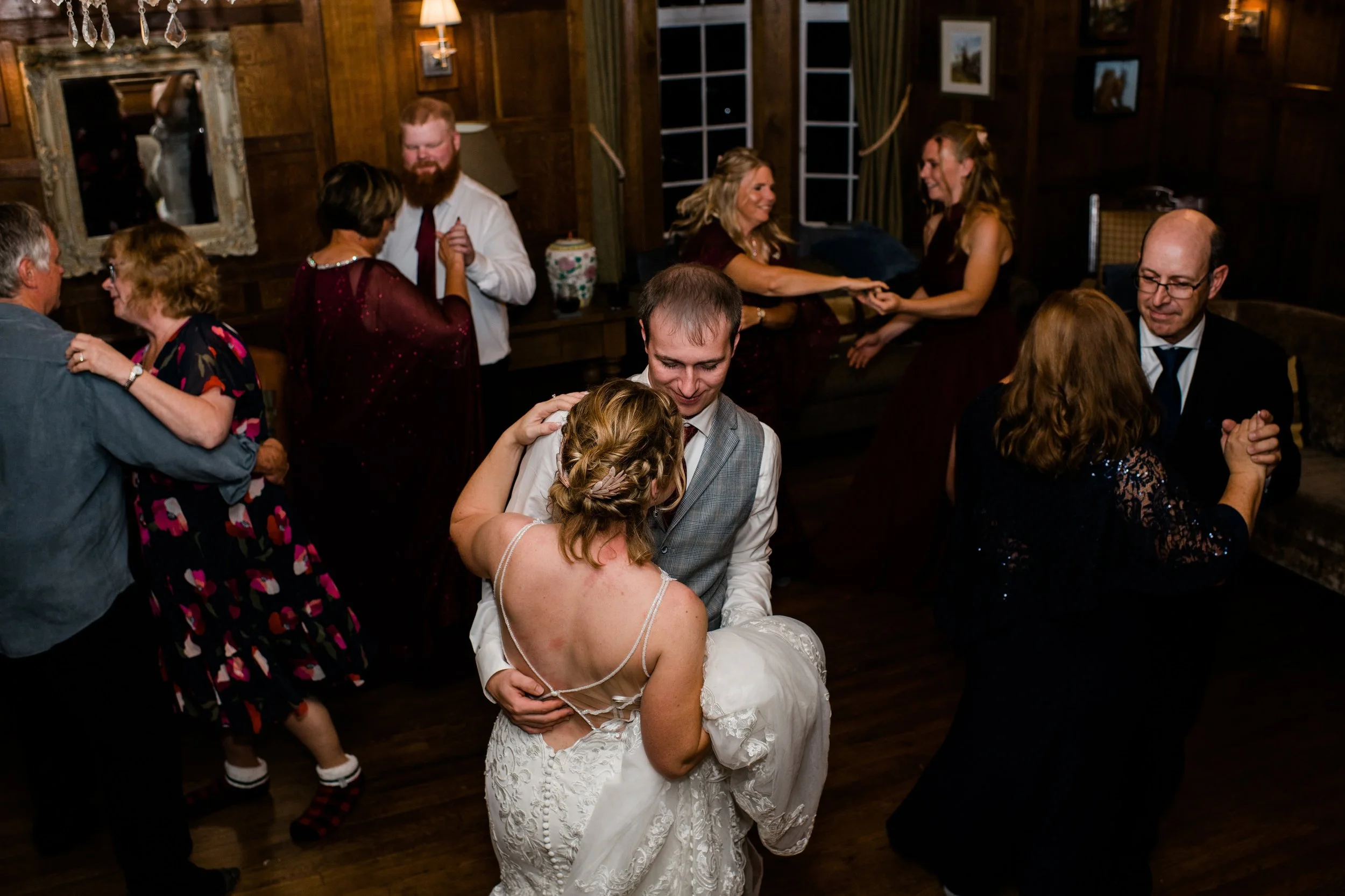 Couple dancing with the woman in a white wedding dress and the man in a vest, surrounded by other couples dancing at a wedding reception in a wood-paneled room.