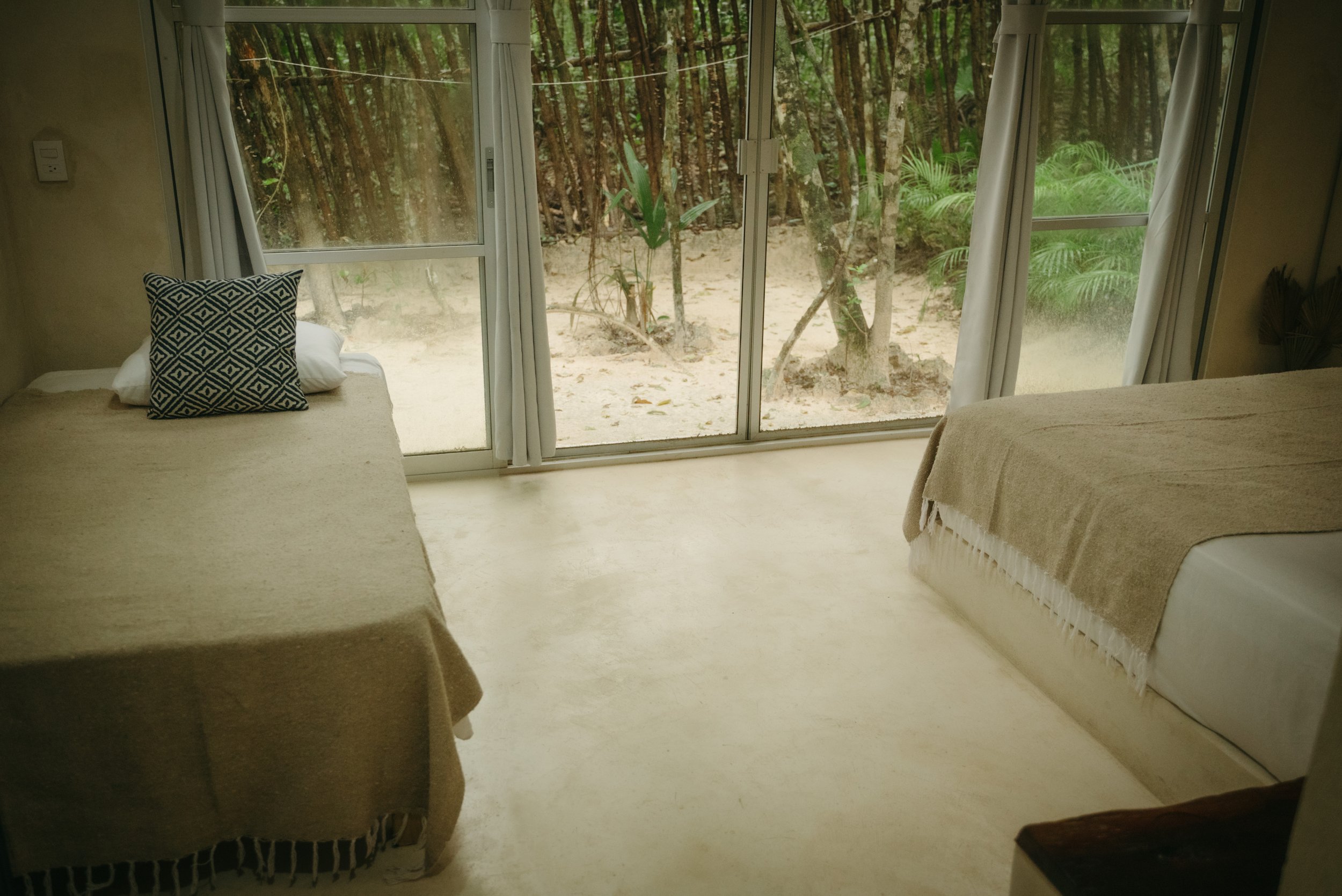 Two beds with beige bedspreads in a room with large glass windows overlooking a sandy outdoor area and trees.