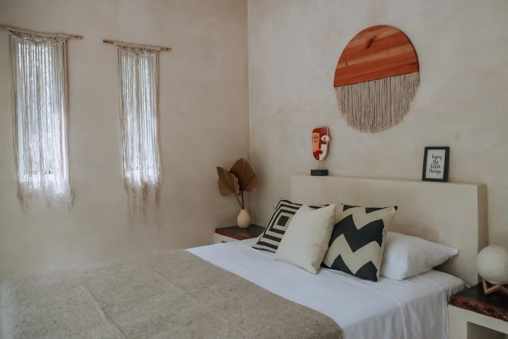 A cozy bedroom with a white bed, decorative pillows, and wall art including a round woven piece and a framed quote. Two windows with sheer curtains let in natural light, with small side tables and decorative dried leaves.