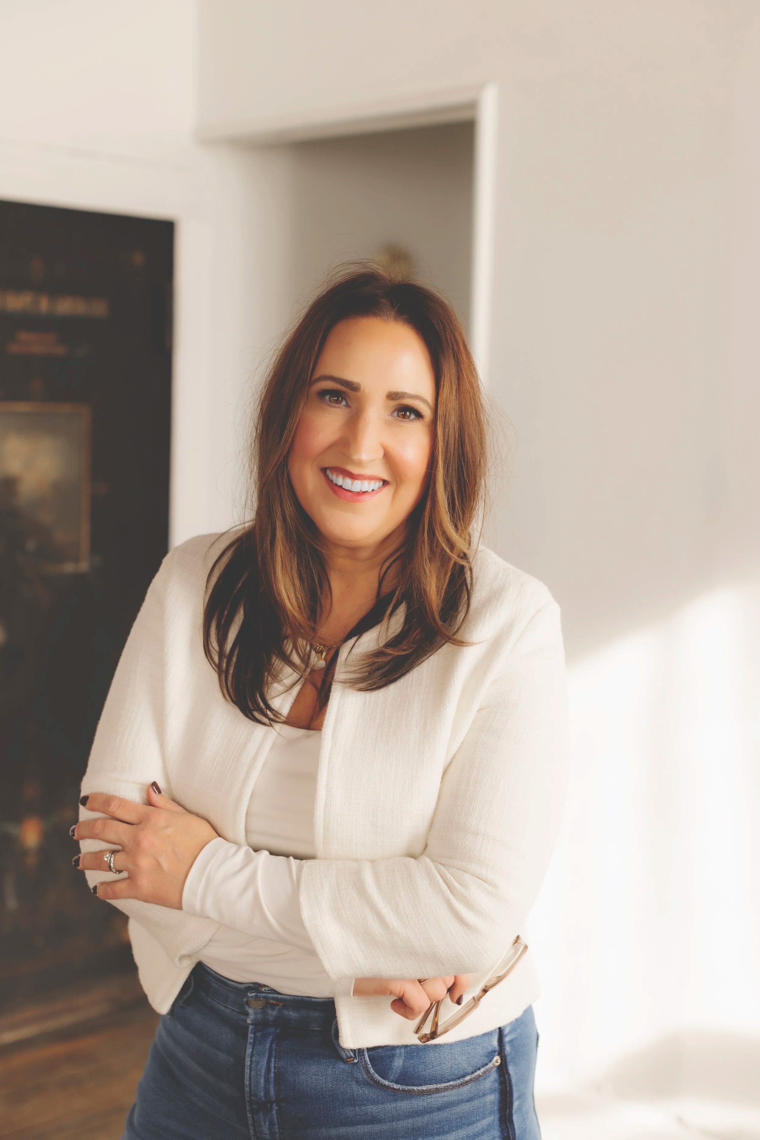 Image of Katie Richburg, a woman with brown hair standing with her arms crossed over her body wearing blue denim jeans and a long sleeve white sweater holding glasses in her hand smiling warmly, depicting an online OCD therapist.