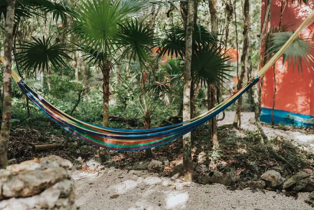 Colorful striped hammock hanging between trees in a tropical forest with dense green foliage and partially visible orange and blue structure in the background.