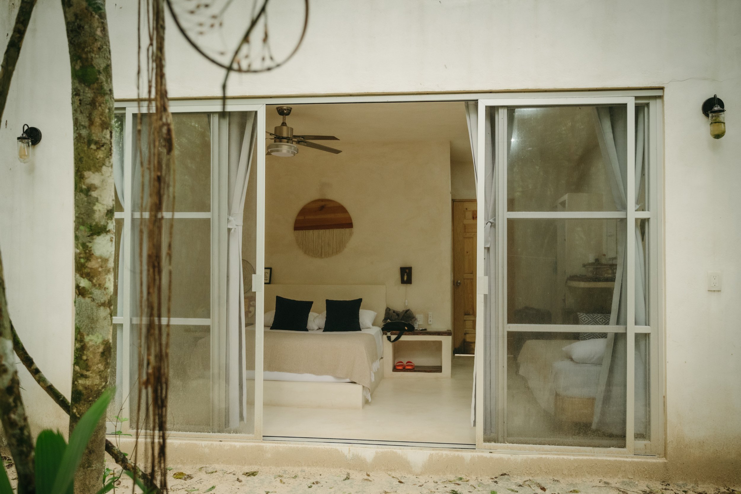 A bedroom with a bed, pillows, and nightstand seen through open glass sliding doors, with a ceiling fan and decor inside.