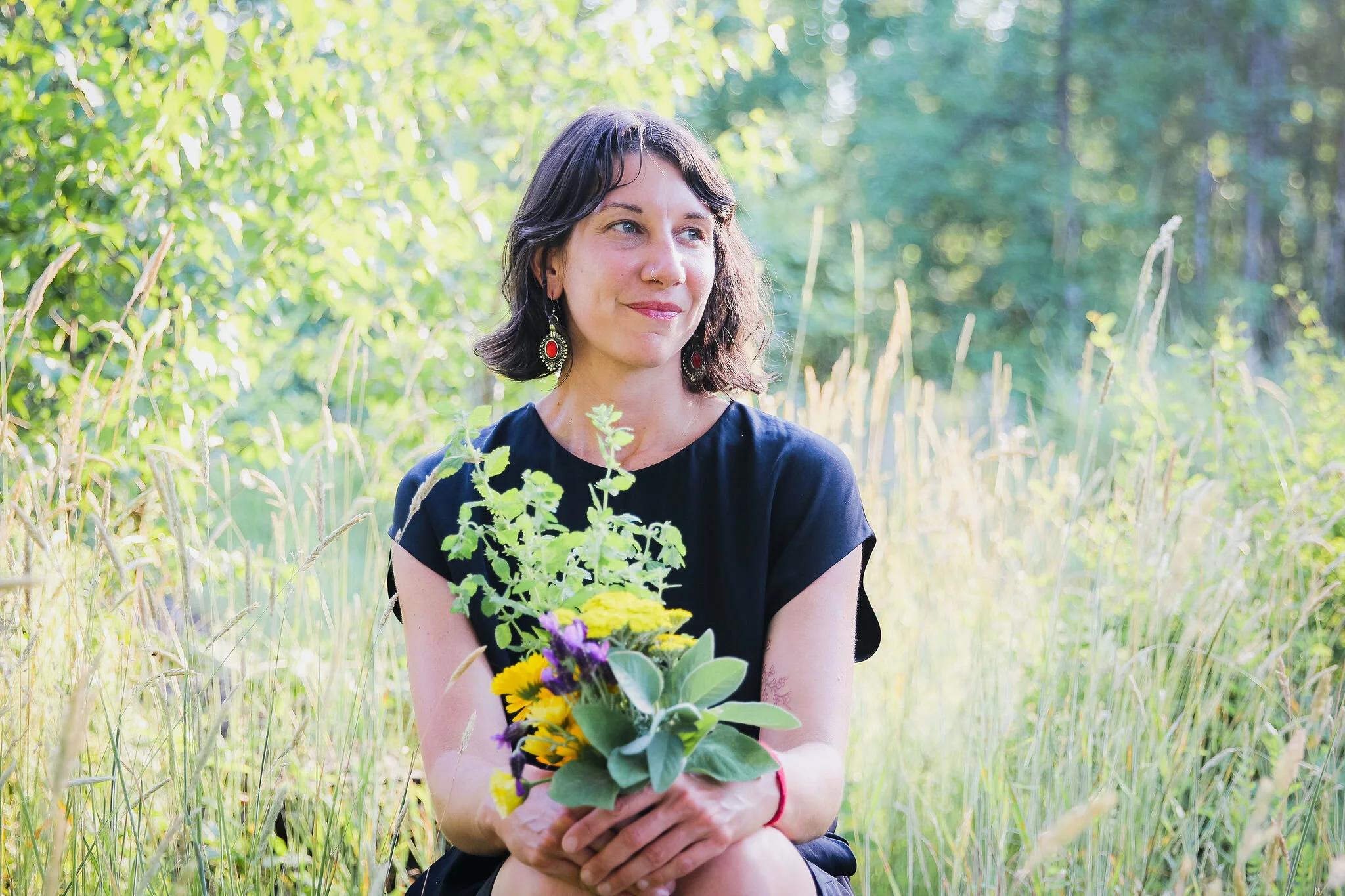 A woman with dark hair and earrings holding a bouquet of colorful flowers while sitting outdoors in a grassy area with green trees in the background.