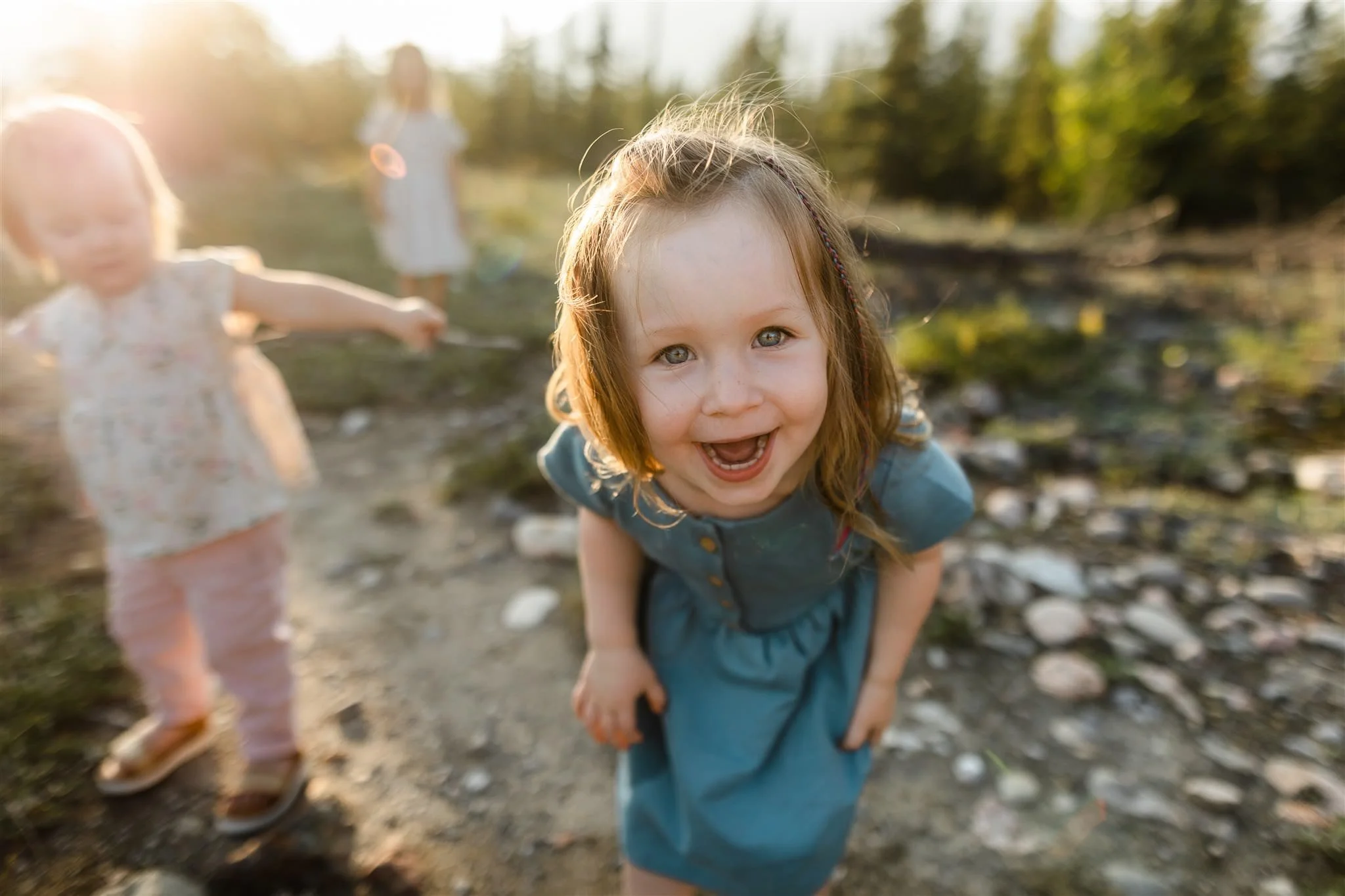 A young girl with brown hair and blue eyes smiling and looking at the camera, outdoors on a sunny day, with two other children blurred in the background.