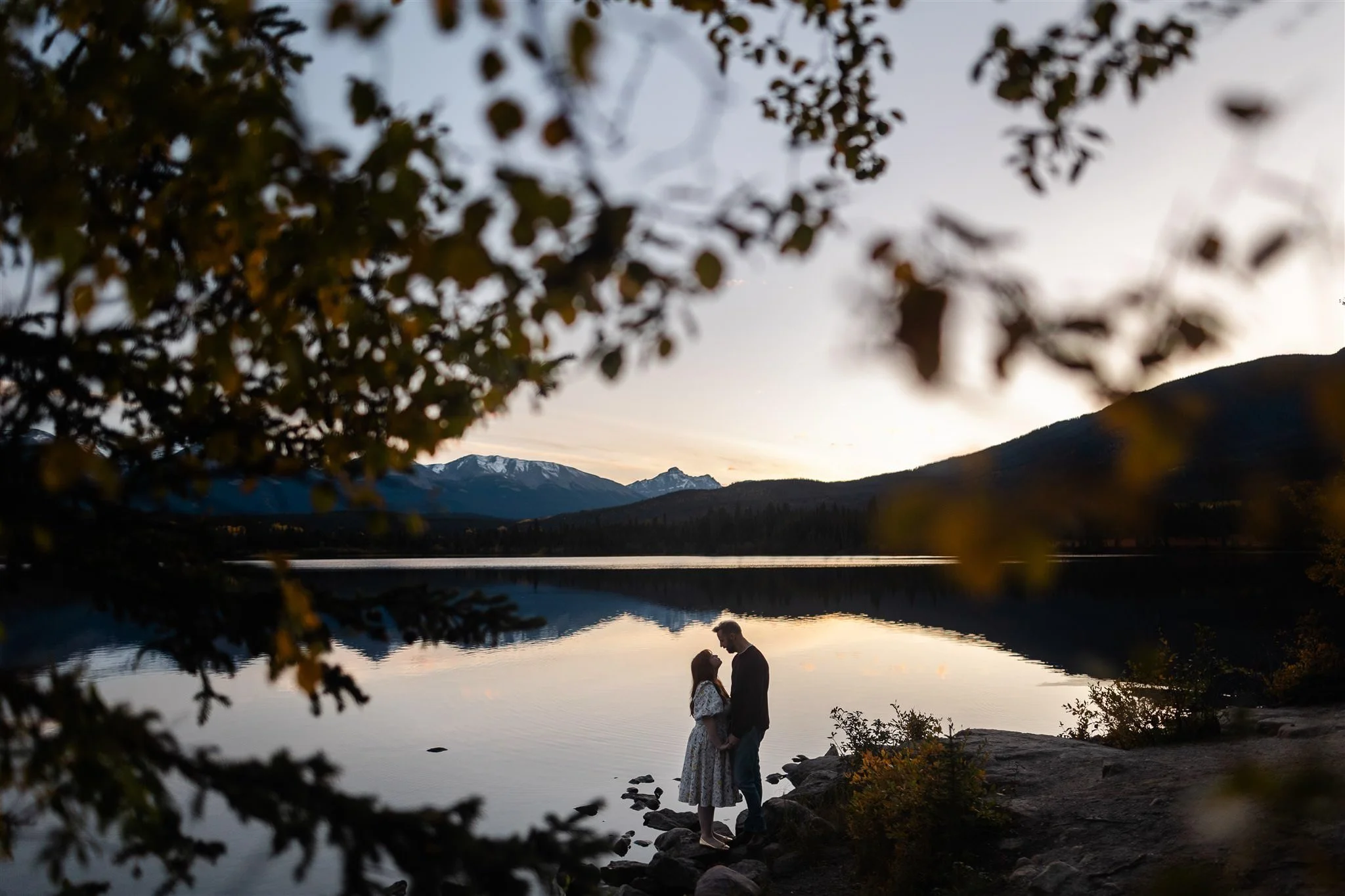 Couple standing by Pyramid lake at sunset with mountains in the background, framed by blurred trees in the foreground.