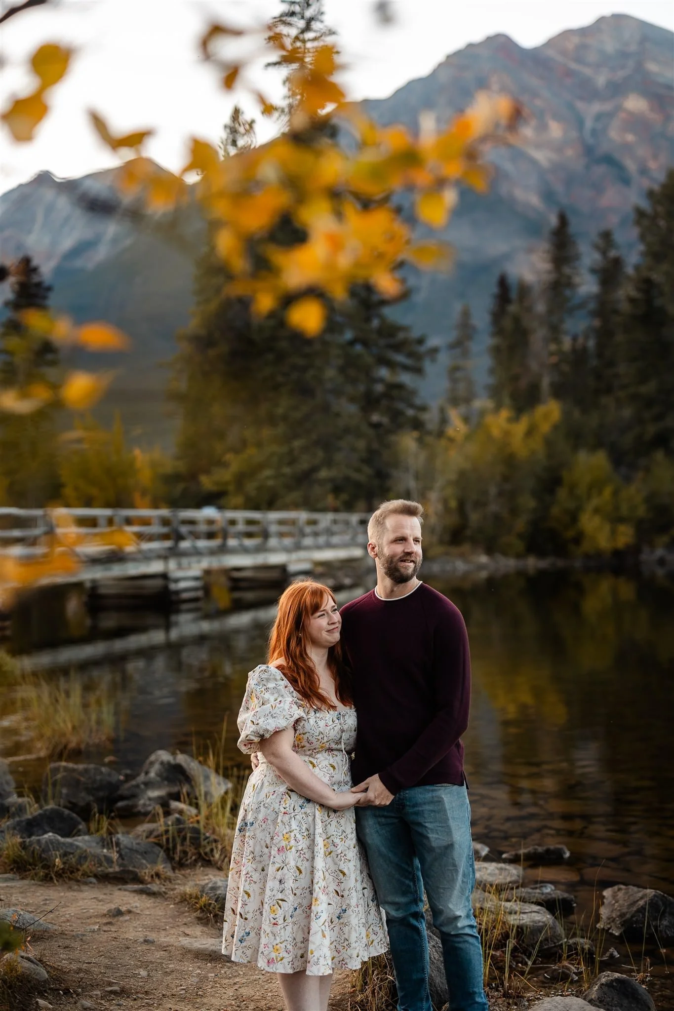 A couple holding hands by a lake with mountains and trees in the background during autumn.