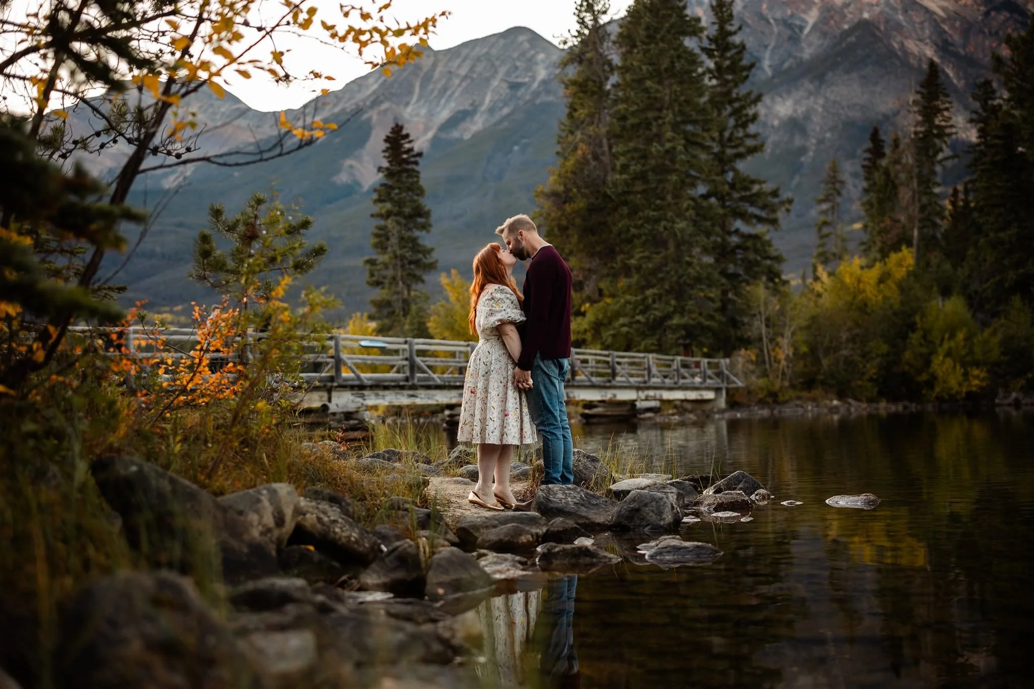 A couple holding hands and touching foreheads by a lake with mountain and forest background, during sunset.