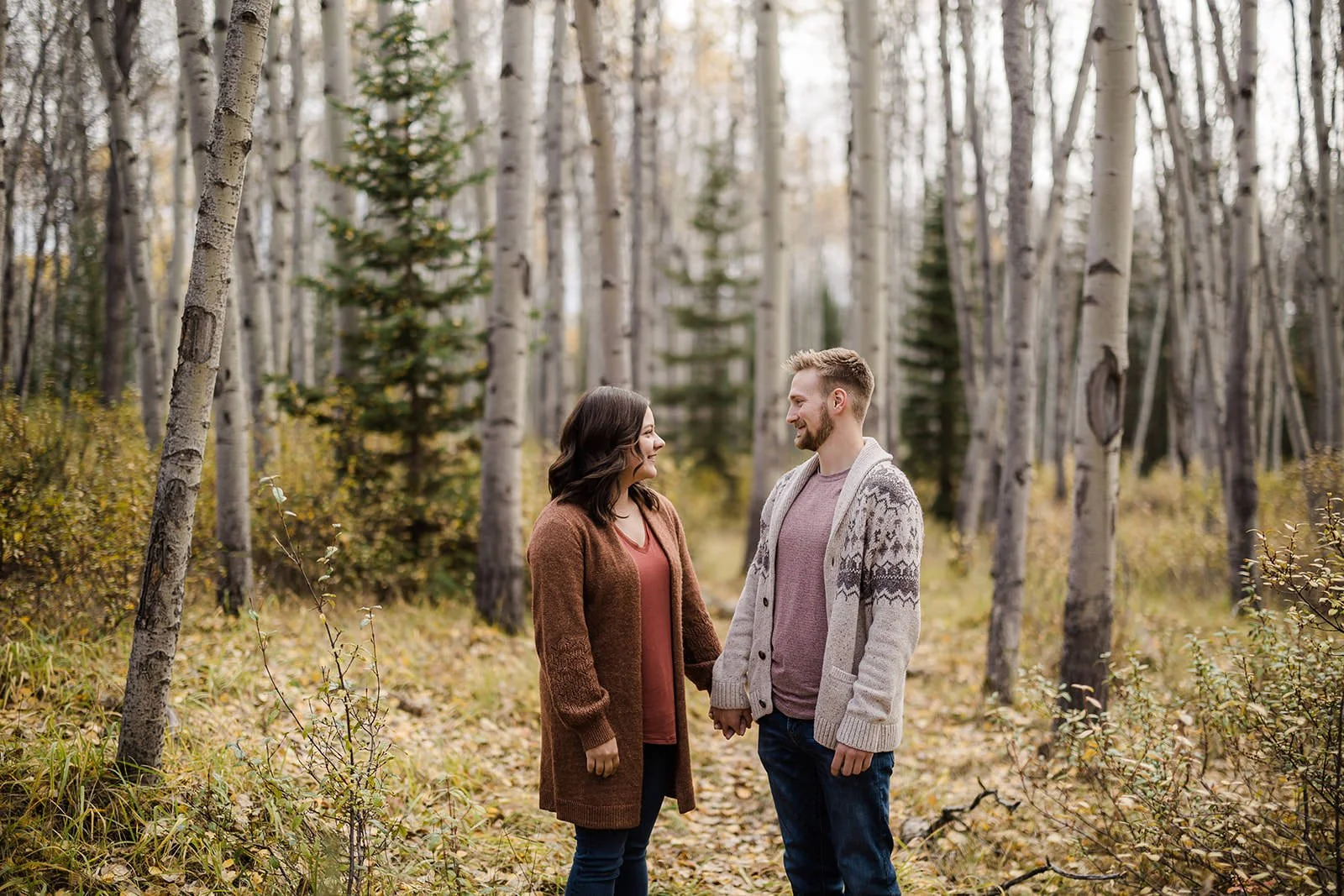 A man and woman standing in a forest, facing each other and holding hands, surrounded by trees and autumn foliage.