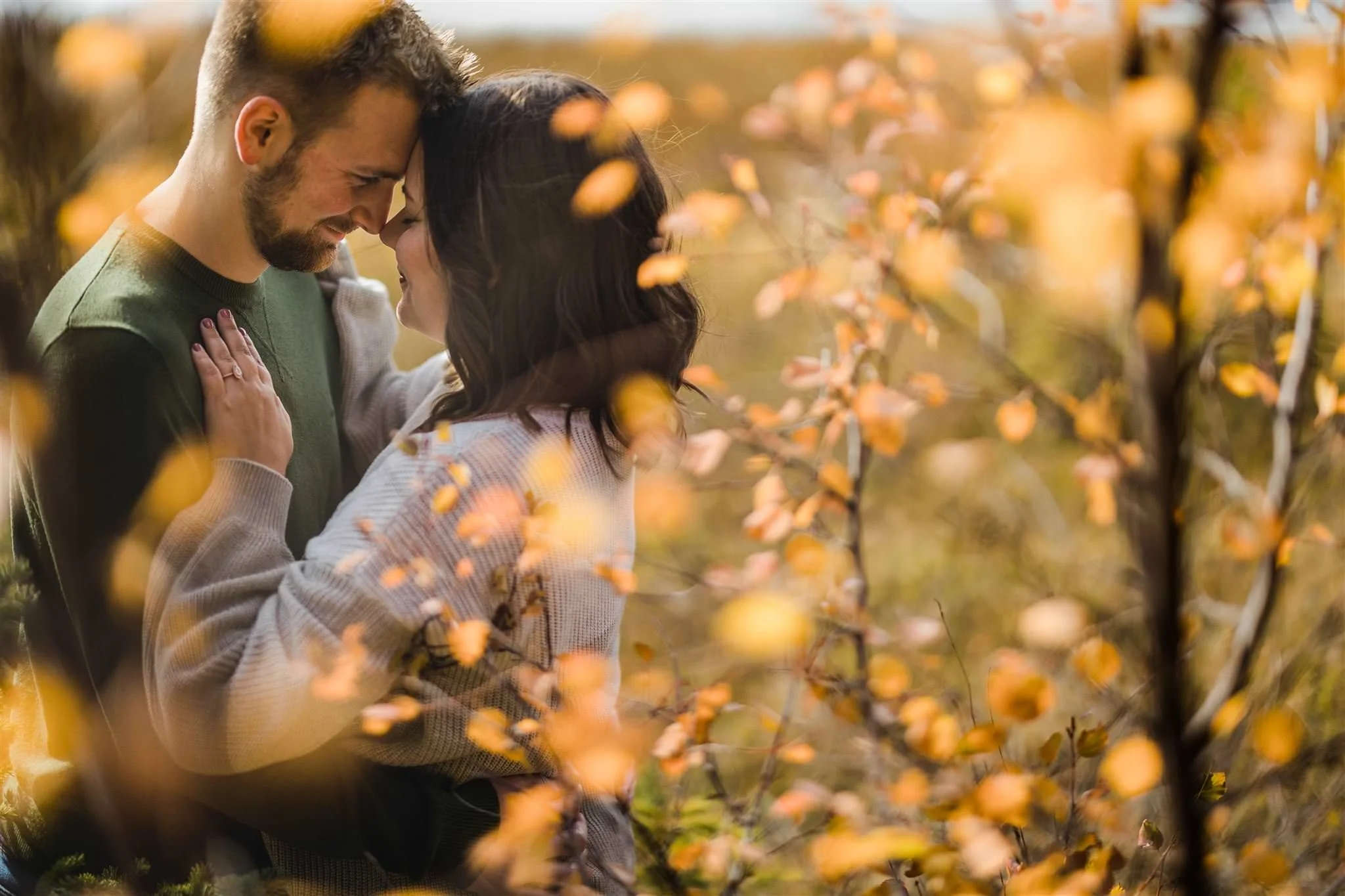 A couple shares an intimate moment in a park, touching foreheads and smiling amidst autumn foliage.