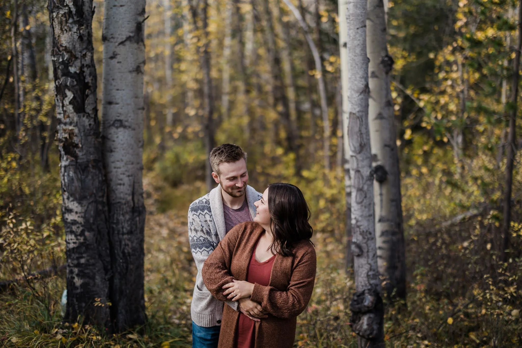 A couple standing close together in a forest with autumn-colored leaves, smiling and looking at each other.