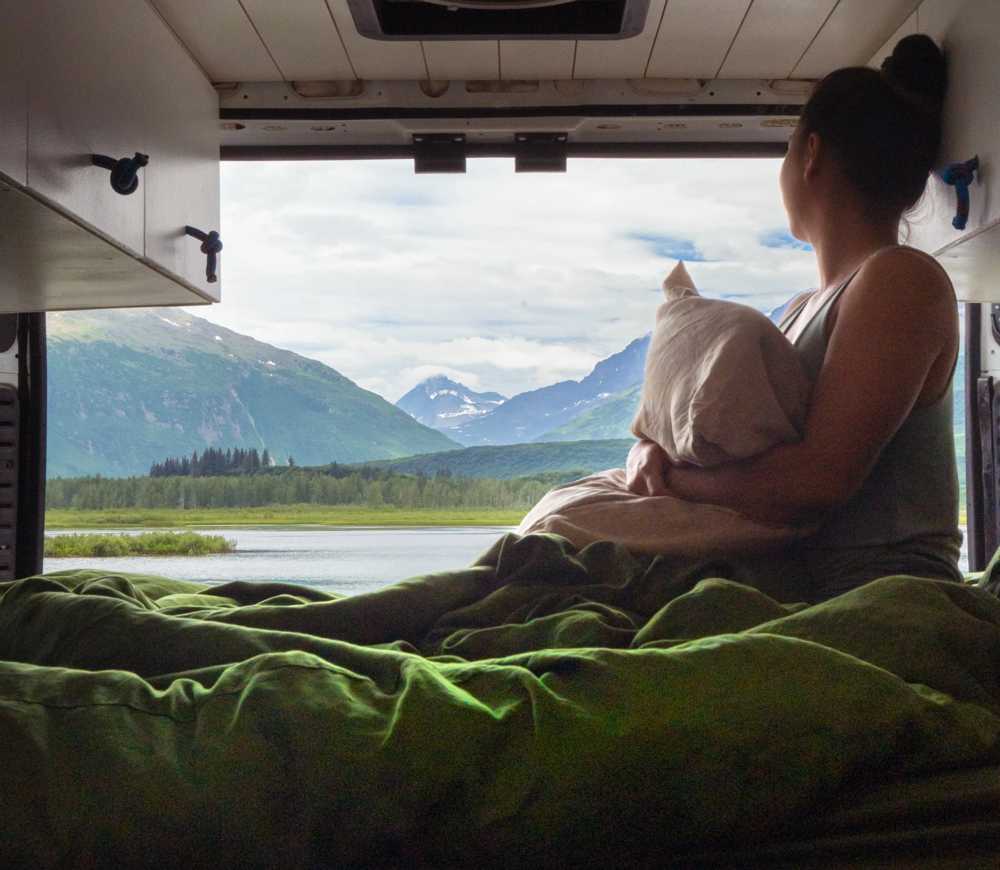 A woman lounging in bed looking over a beautiful lake and mountain scene in Alaska.