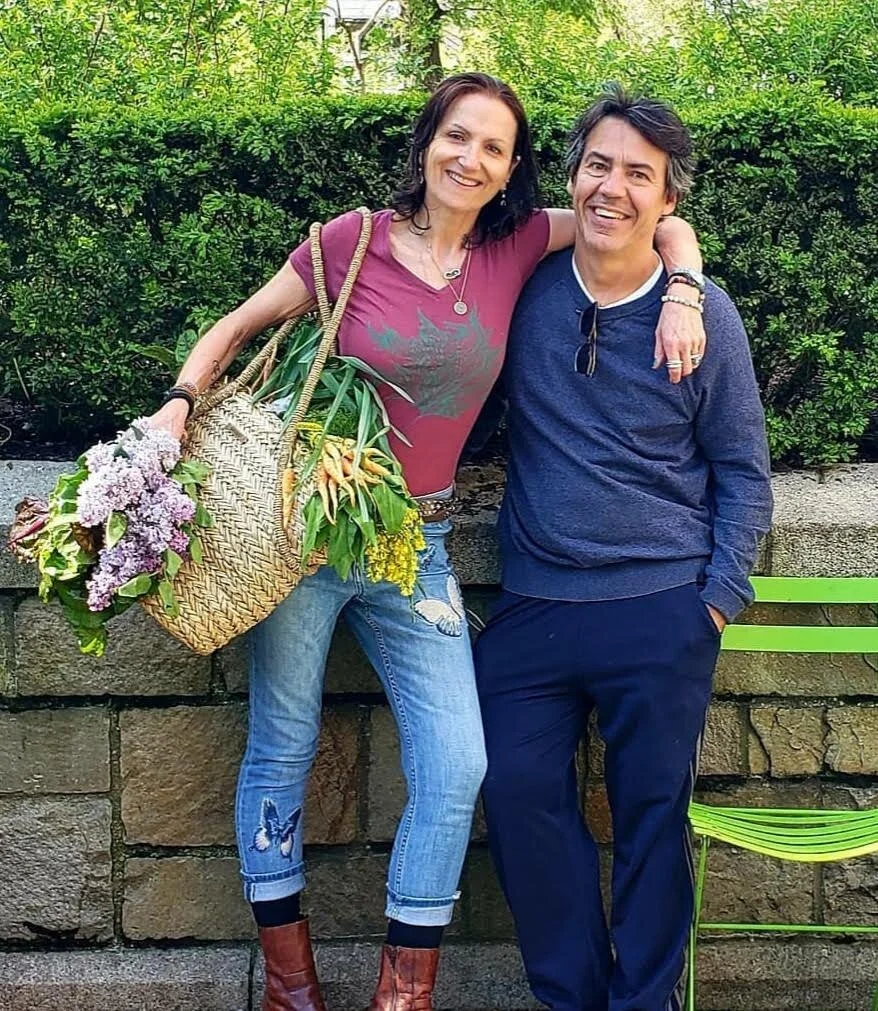 Two people smiling outdoors; one holding a basket of flowers and vegetables.