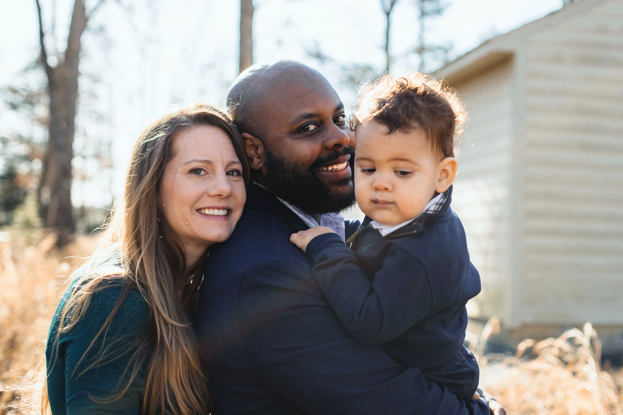 A family portrait with a smiling woman, a man, and a young child outside in a natural setting.