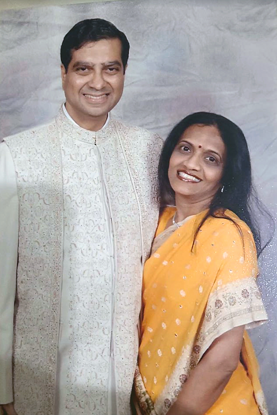 A smiling couple dressed in traditional Indian attire; the man is wearing an embroidered sherwani, and the woman is wearing a yellow sari with a patterned border, against a textured background.