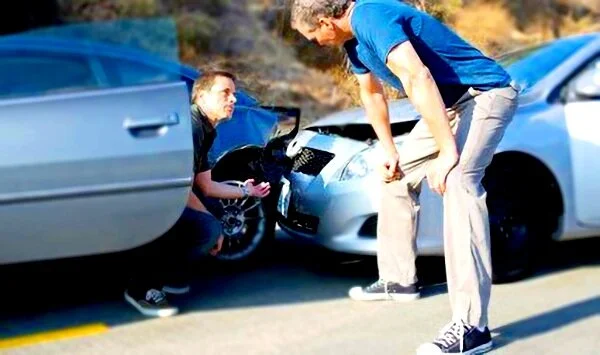 Two men examining damage after a car accident, inspecting the front of two colliding vehicles on a road.