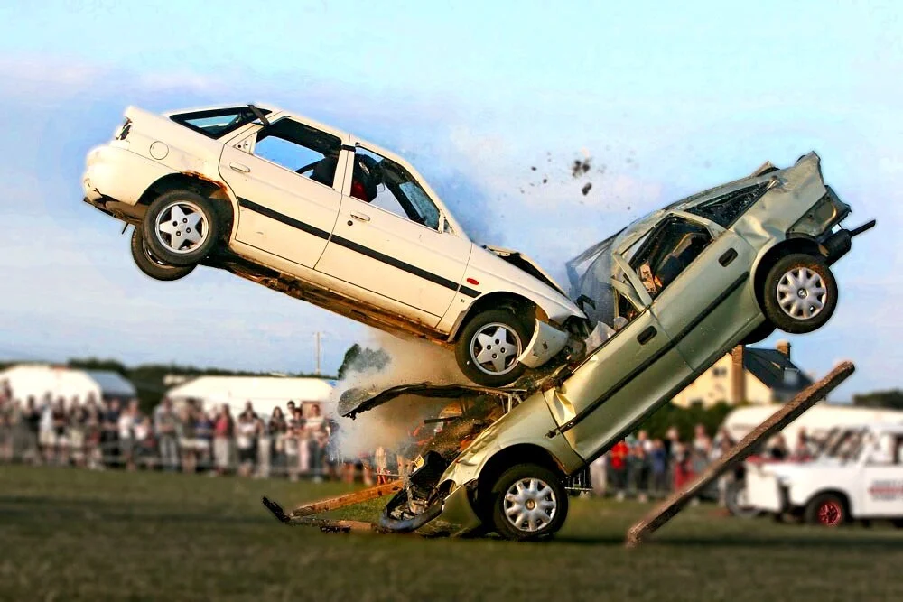Two cars crashing in mid-air at a demolition derby event with spectators in the background.