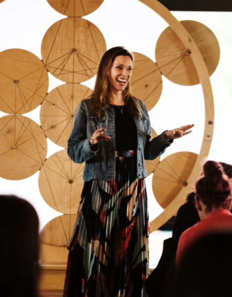 Woman speaking in front of wooden geometric design, wearing denim jacket and colorful skirt, addressing seated audience.