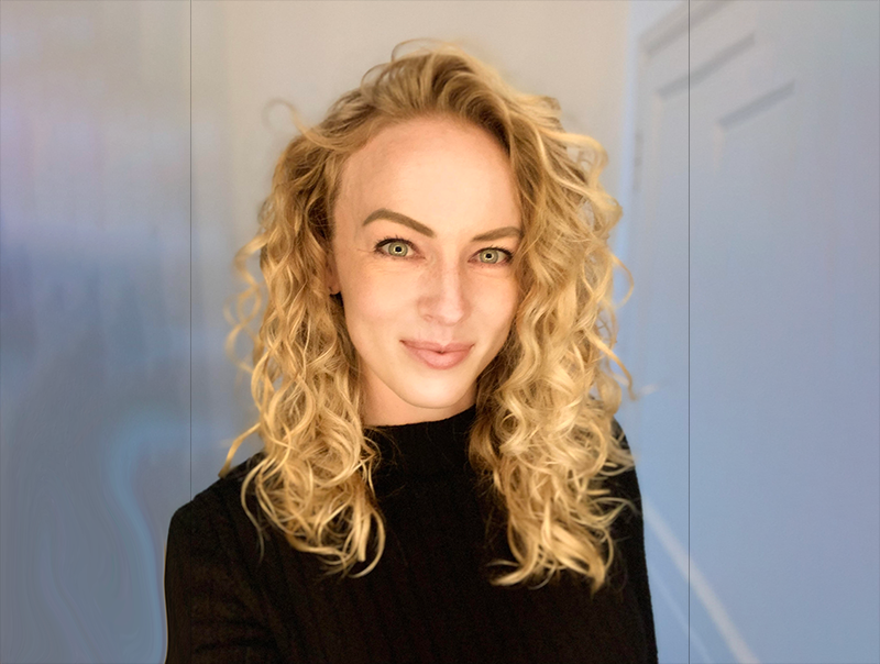Person with curly blonde hair wearing a black shirt, smiling in a bright setting.