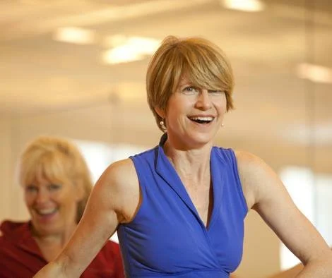 Two women enjoying a fitness or dance class, one in a blue top smiling and leading, with another in a red top in the background.