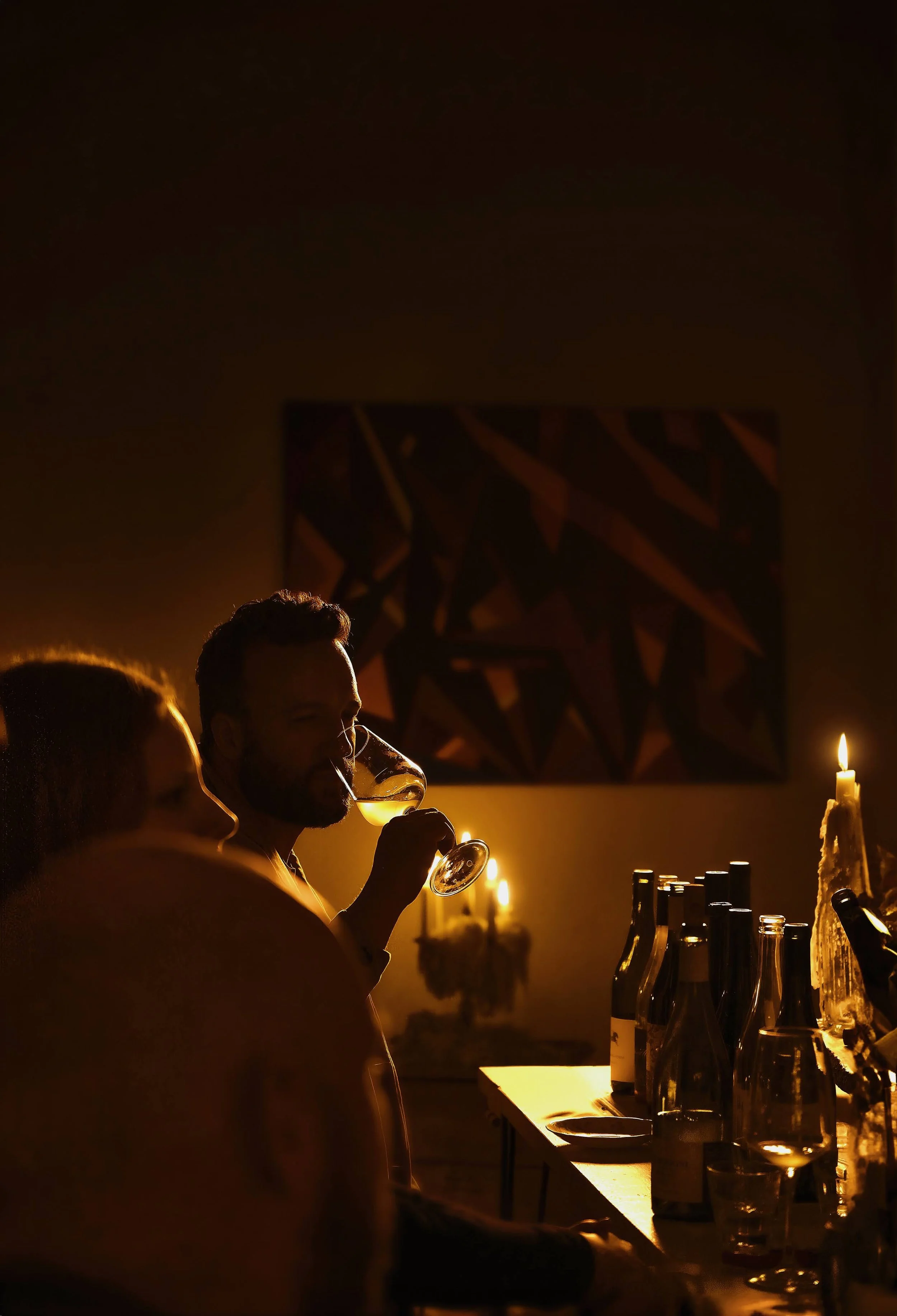 A man and woman sitting at a bar, with the man holding a glass of wine, illuminated by warm yellow lighting and candles, with artwork on the wall behind them.