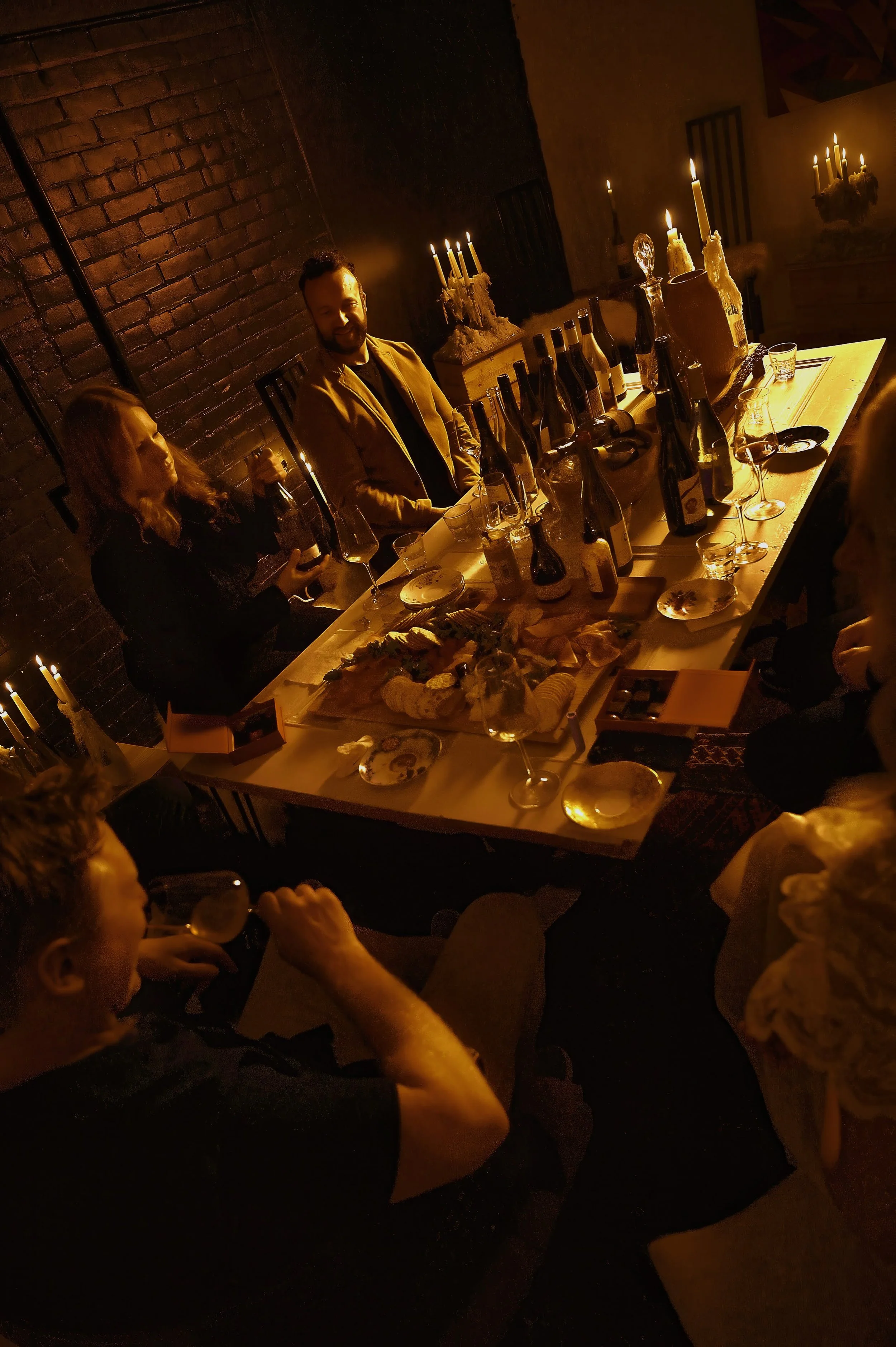 A group of people sitting around a dinner table in a dimly lit room with brick walls and candles, enjoying drinks and snacks.