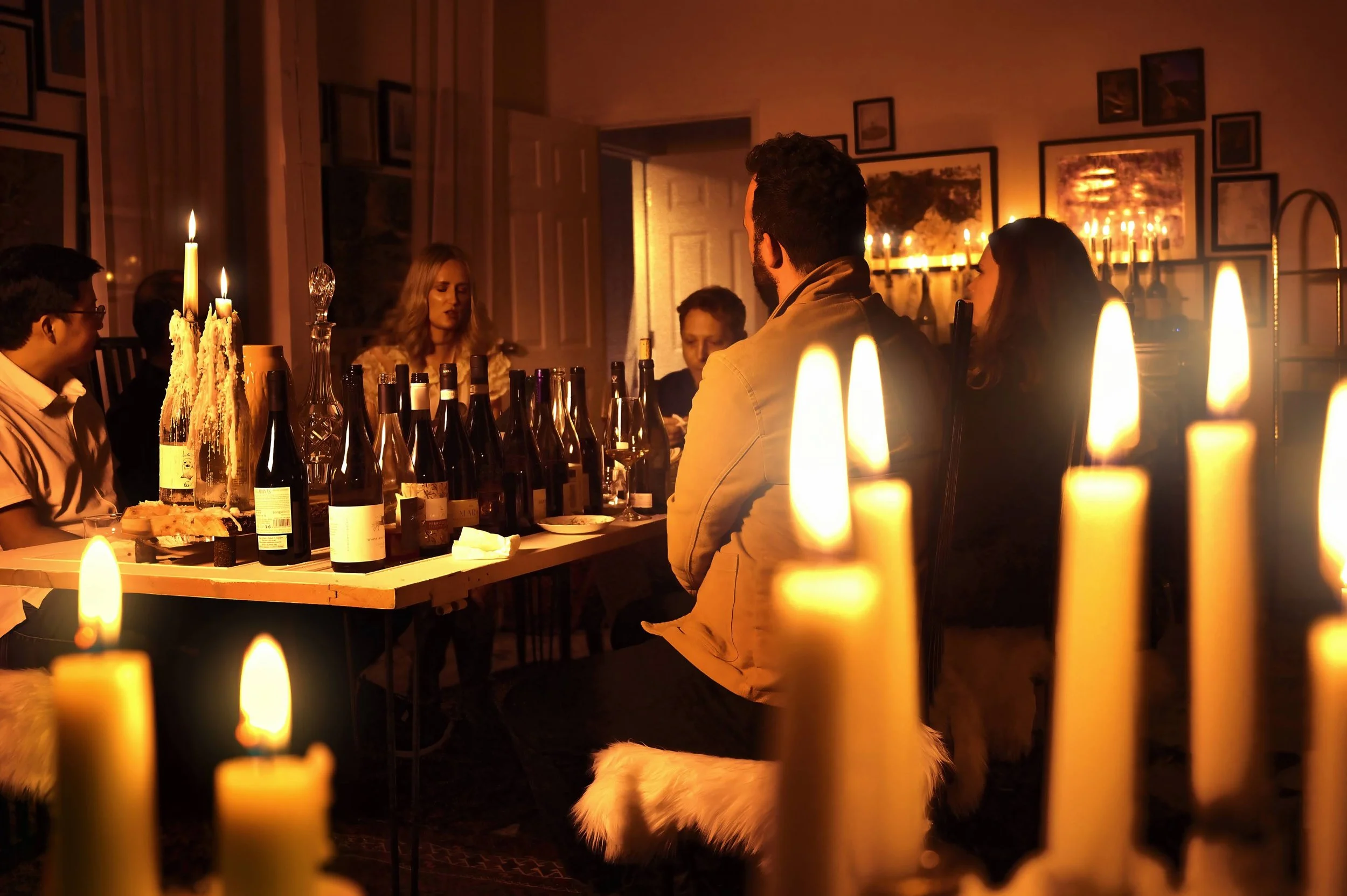 People gathered around a dining table in a dimly lit room with candles, bottles of wine, and pictures on the wall.