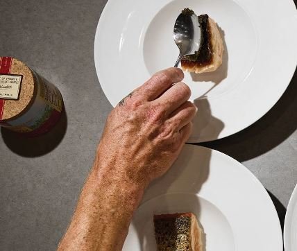 A hand holding a spoon about to eat a slice of chocolate cake on a white plate, with another slice on a different plate and a jar of Nutella nearby.